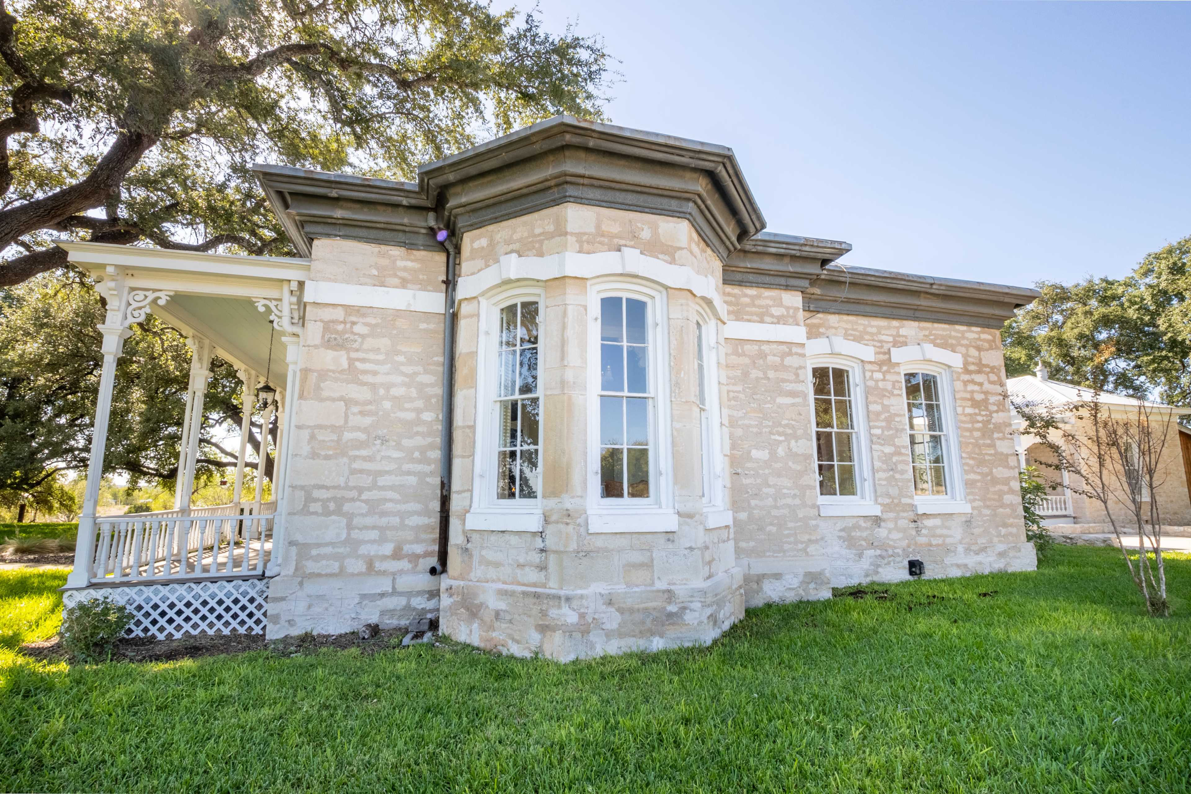 A historic stone house with large windows and a decorative porch, situated on a grassy area.