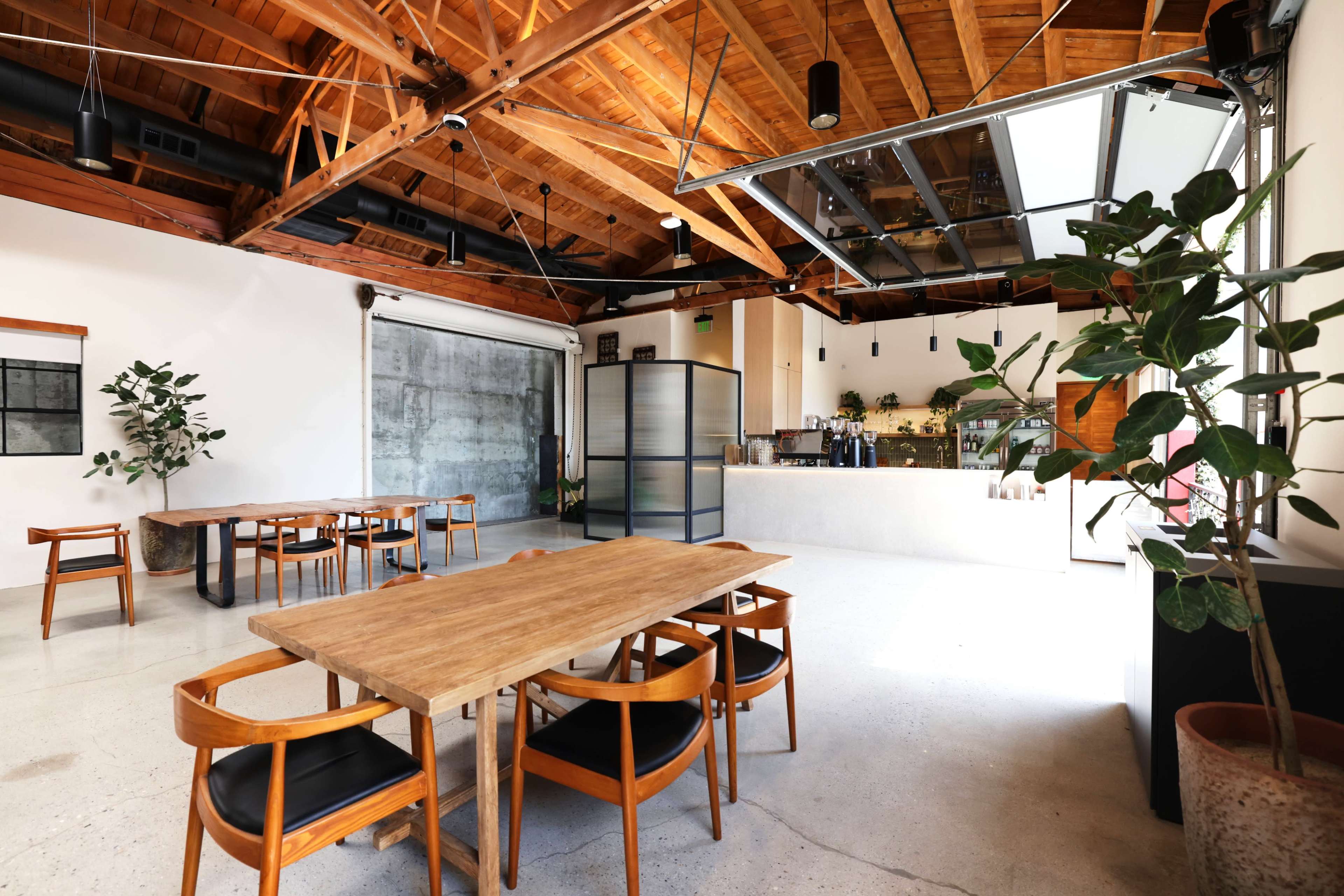 The image shows an open space with a high wooden ceiling, featuring a dining area with a wooden table and chairs, alongside a modern bar setup.