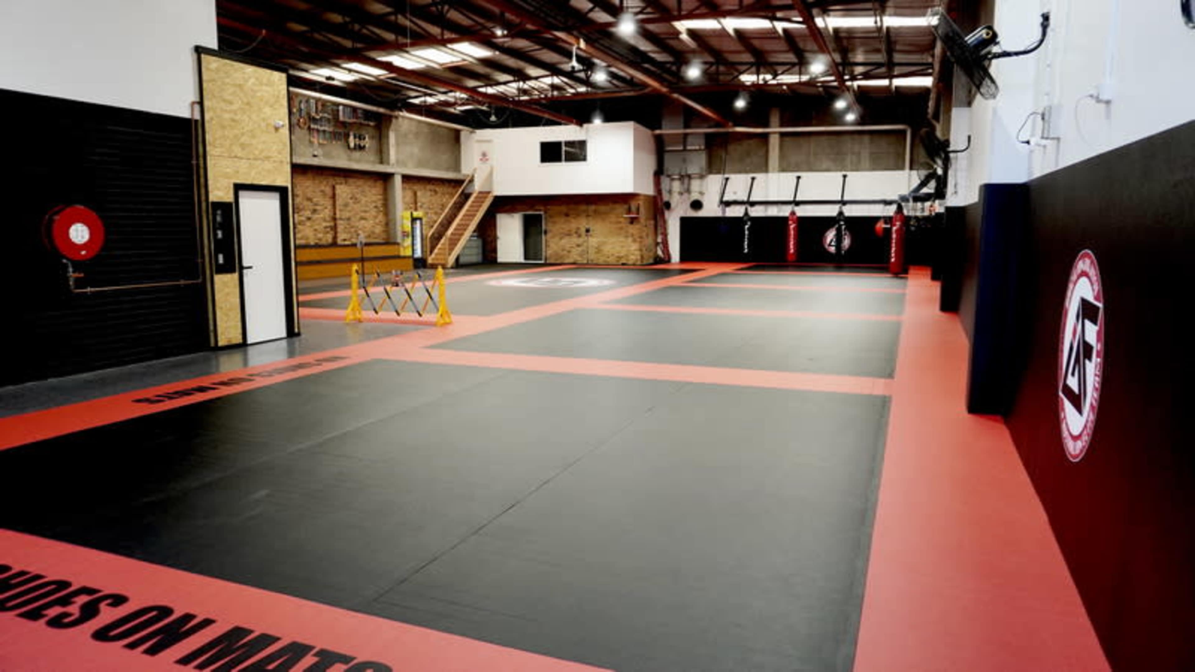 The image shows a spacious training facility with black and red padded mats, framed by brick and concrete walls, along with various training equipment and a staircase in the background.