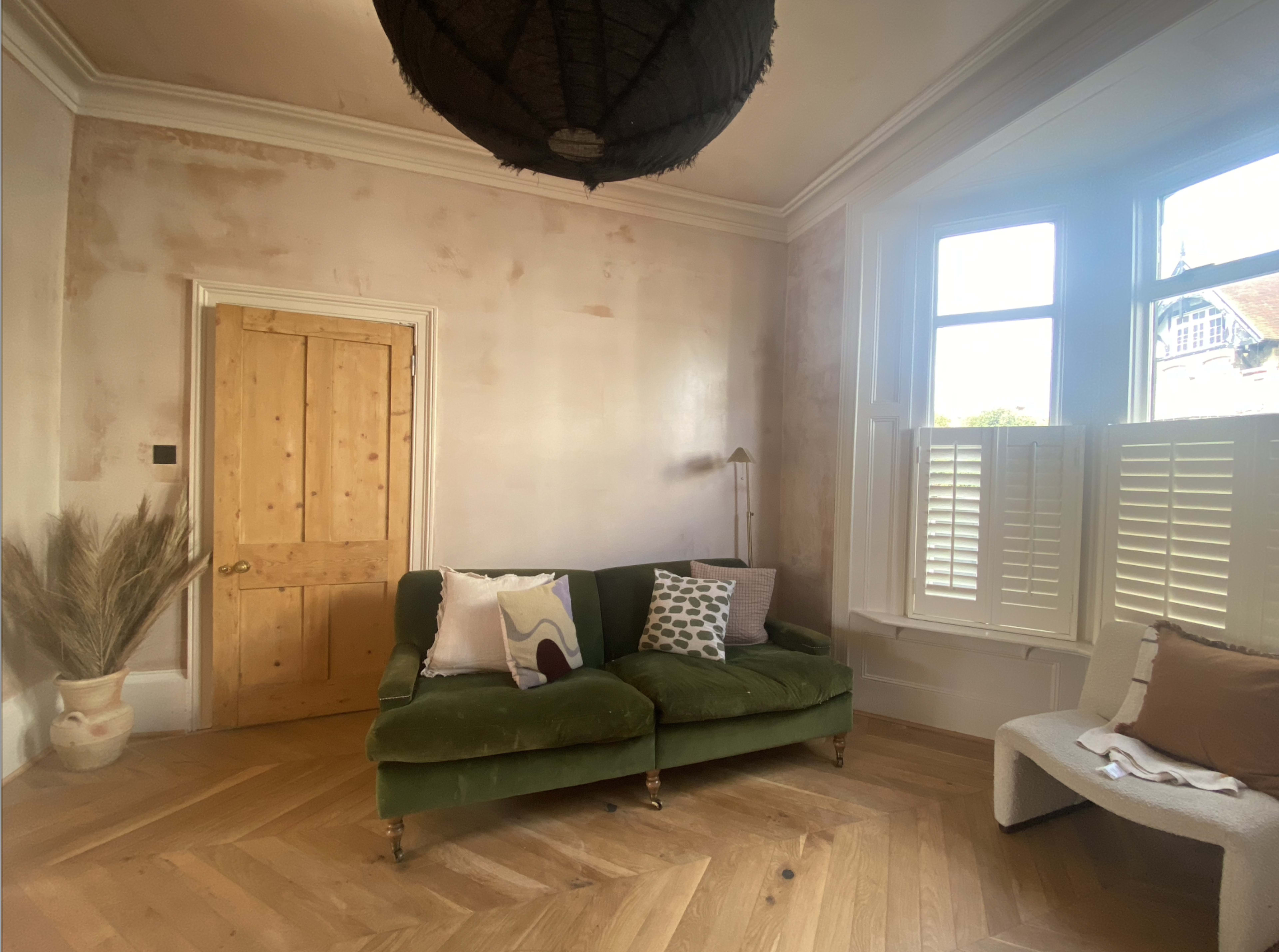 A room with a green sofa, a wooden door, and large windows with shutters, featuring a light-colored floor and decorative items.