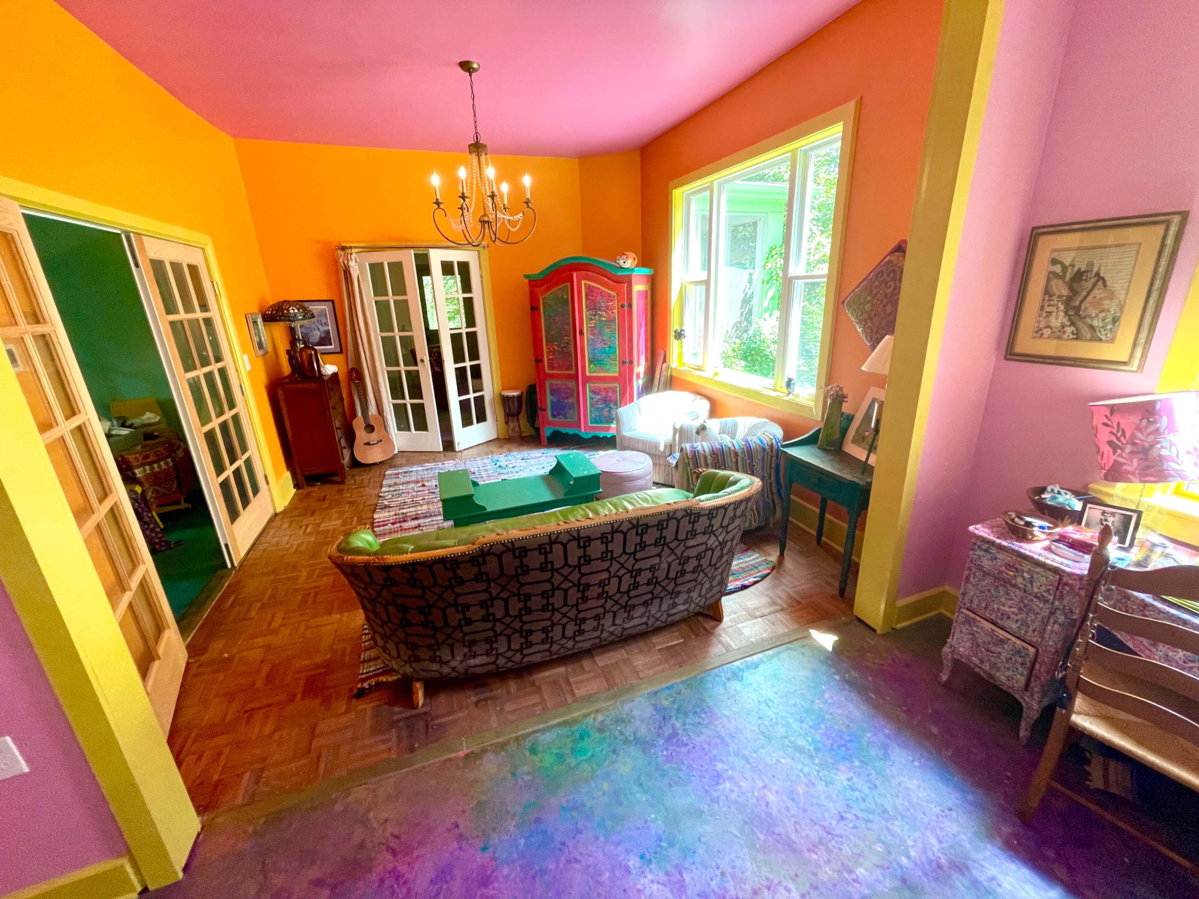 The image shows a brightly colored living room with orange and pink walls, a patterned couch, large windows, and various pieces of furniture.