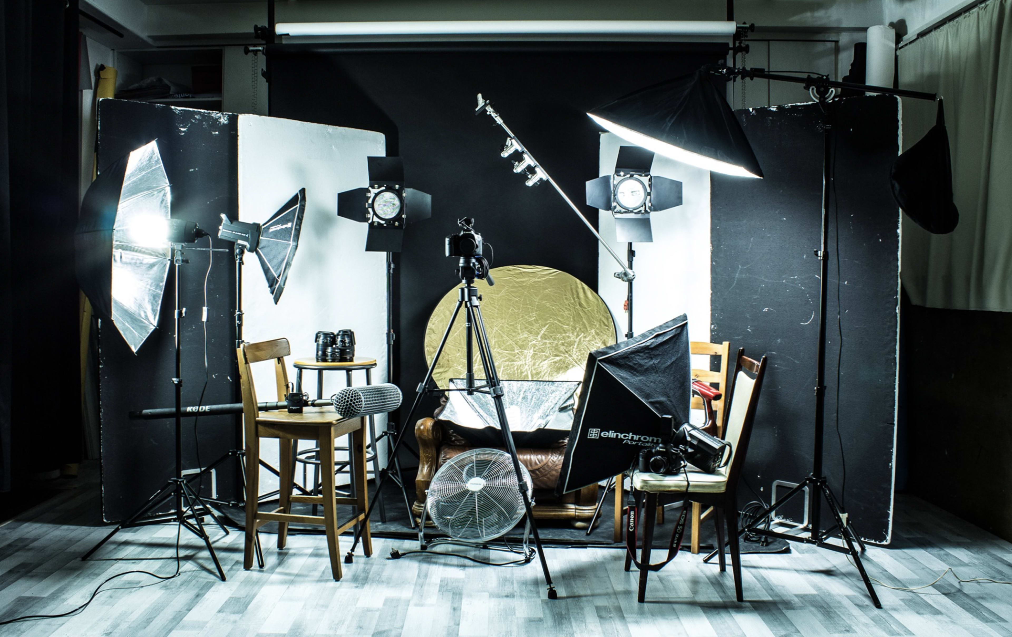 A photography studio setup with various lighting equipment, a camera on a tripod, and multiple chairs positioned around a backdrop.