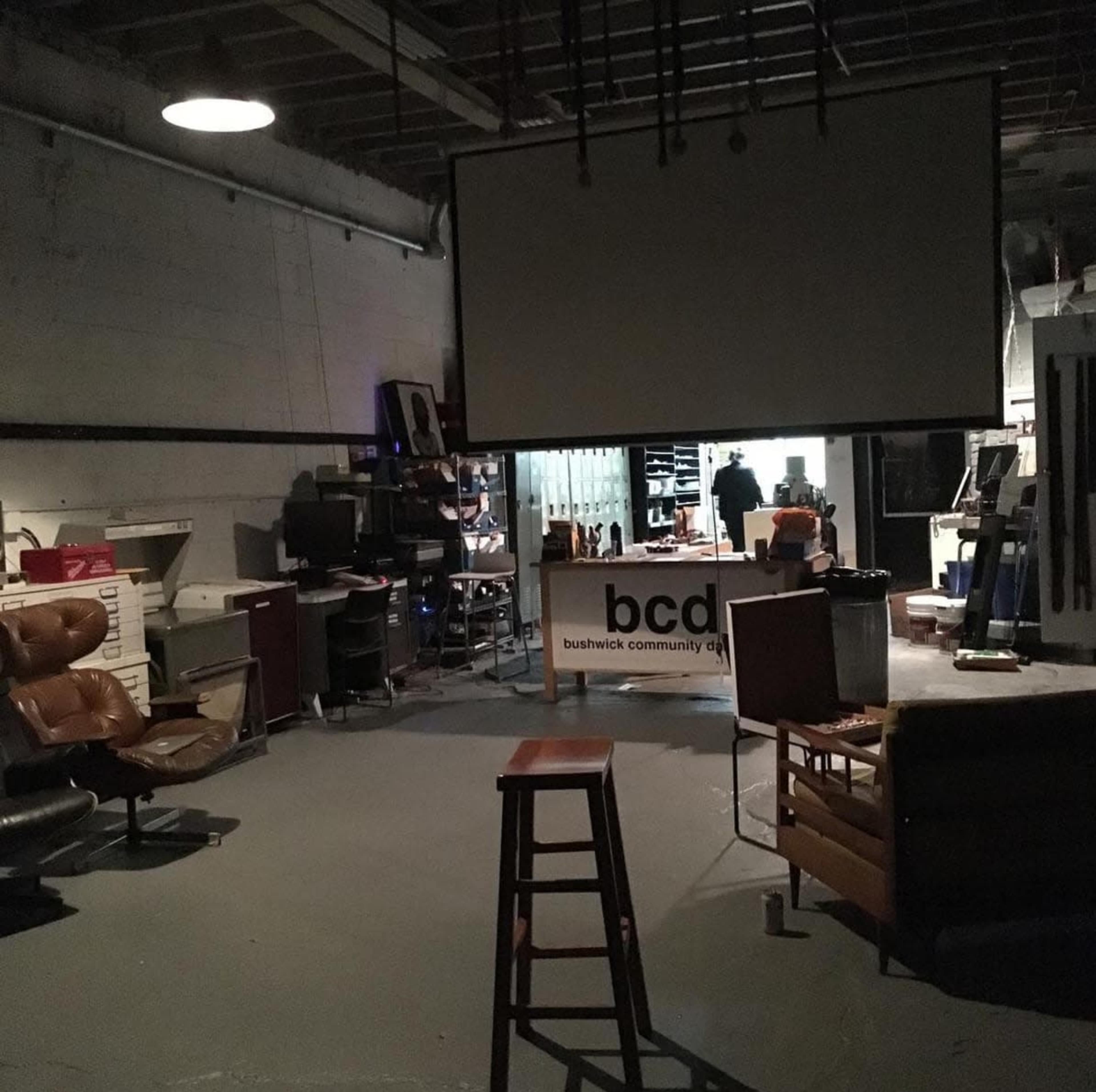 The image shows a dimly lit room with a bar stool in the foreground, surrounded by furniture and office equipment, with a sign for "bcd" in the background.