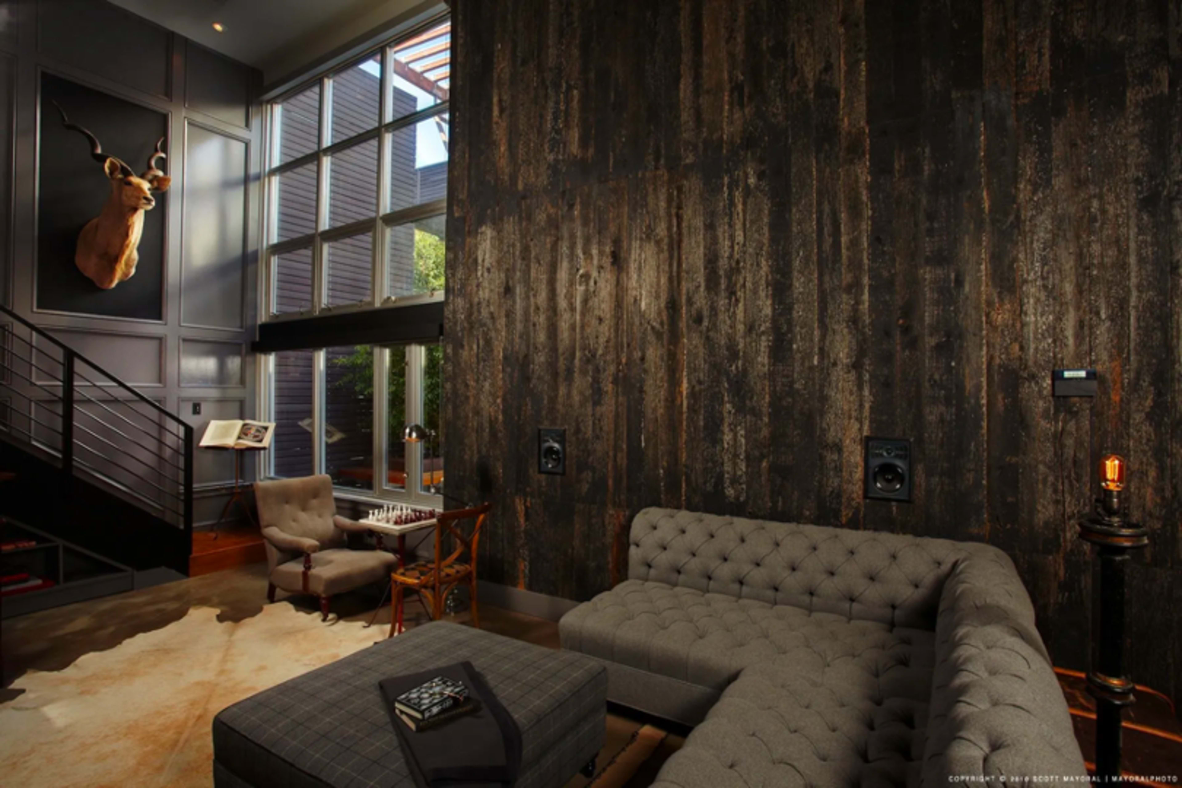 The scene features a stylish living room with a large wooden wall, a mounted deer head, a gray tufted couch, and large windows allowing natural light to enter.