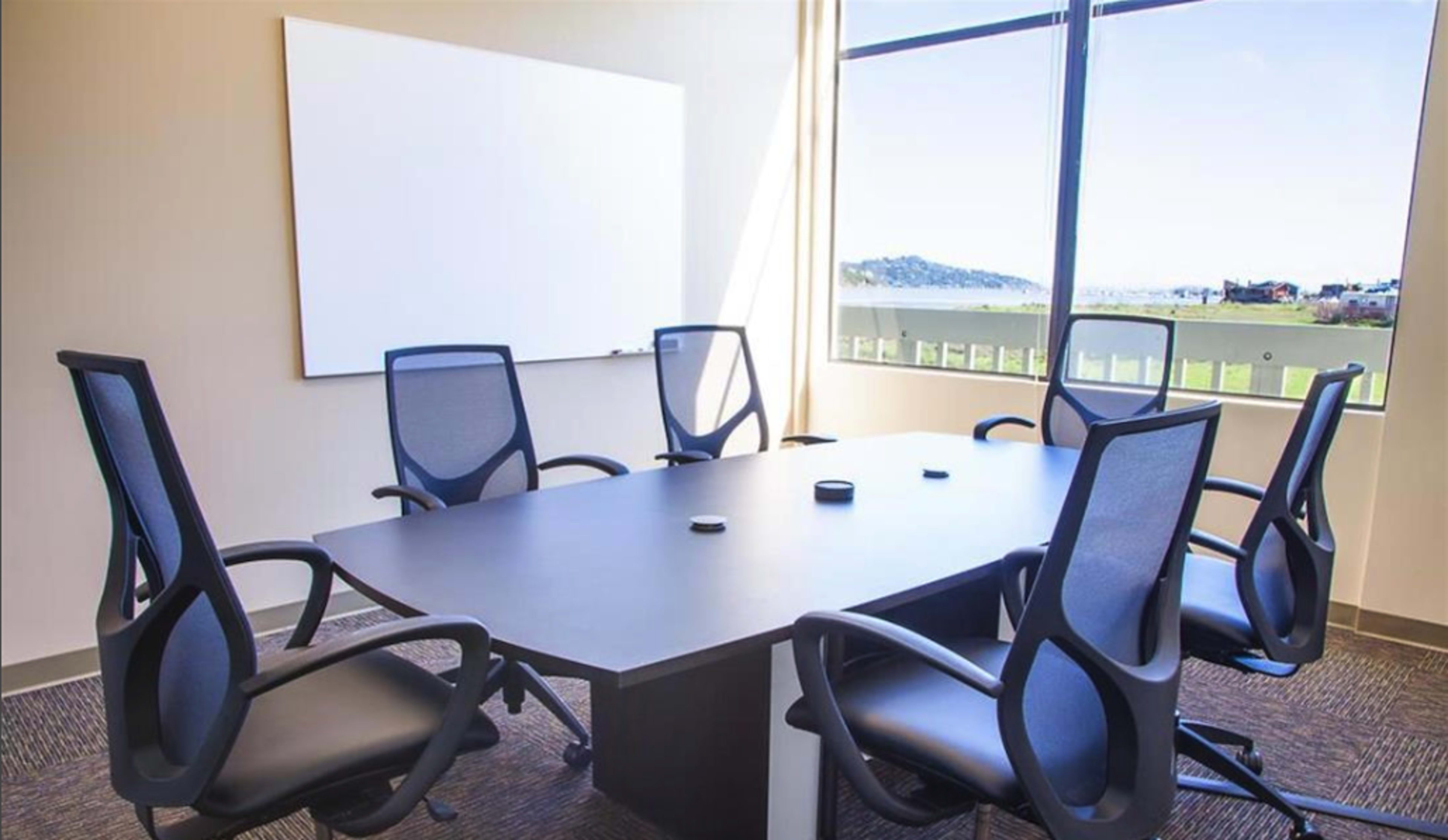 A conference room with a rectangular table surrounded by five black mesh office chairs, a whiteboard on the wall, and large windows letting in natural light.