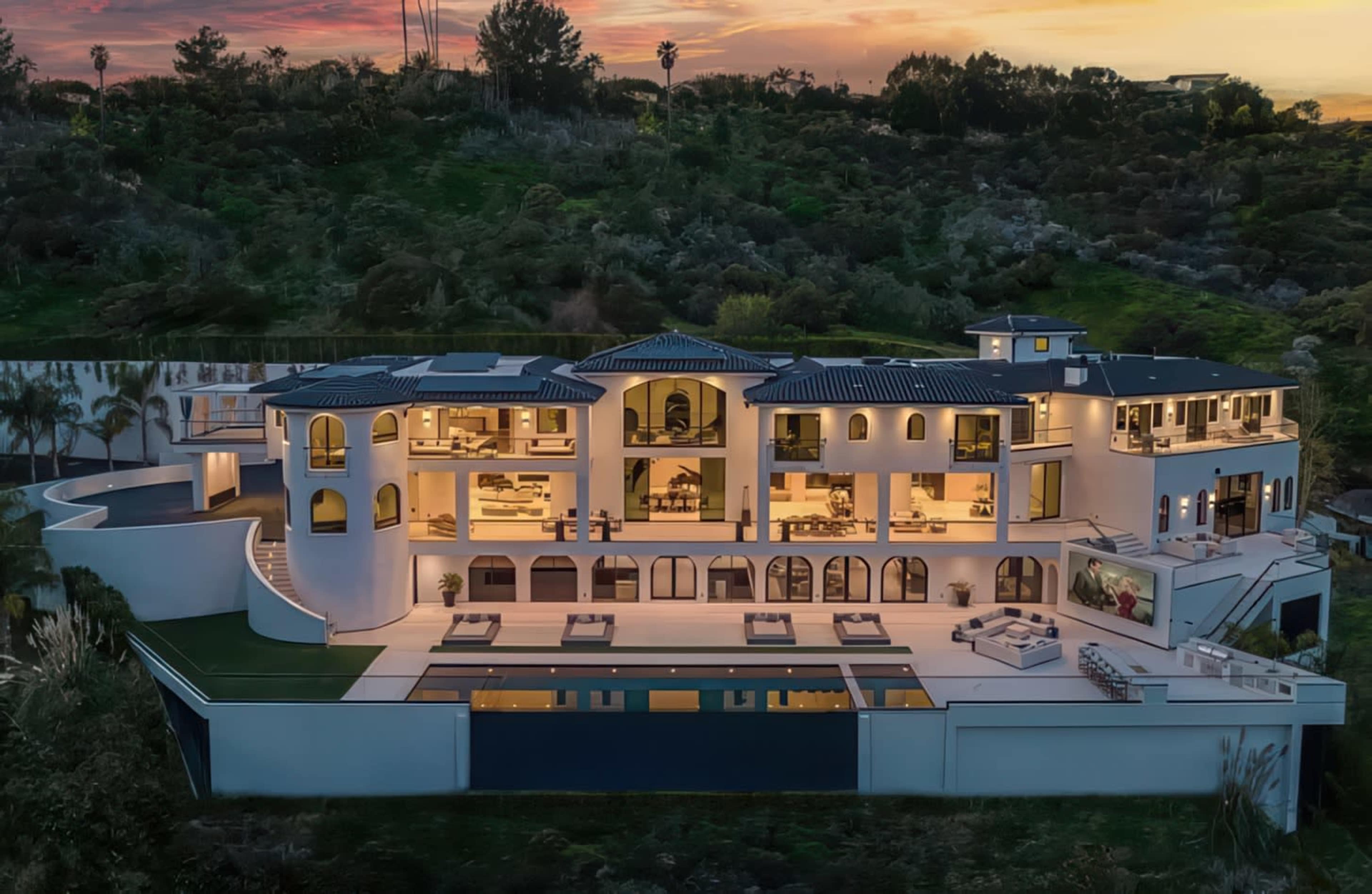 A modern, multi-level luxury mansion with large glass windows and a pool is situated on a hillside surrounded by greenery at sunset.