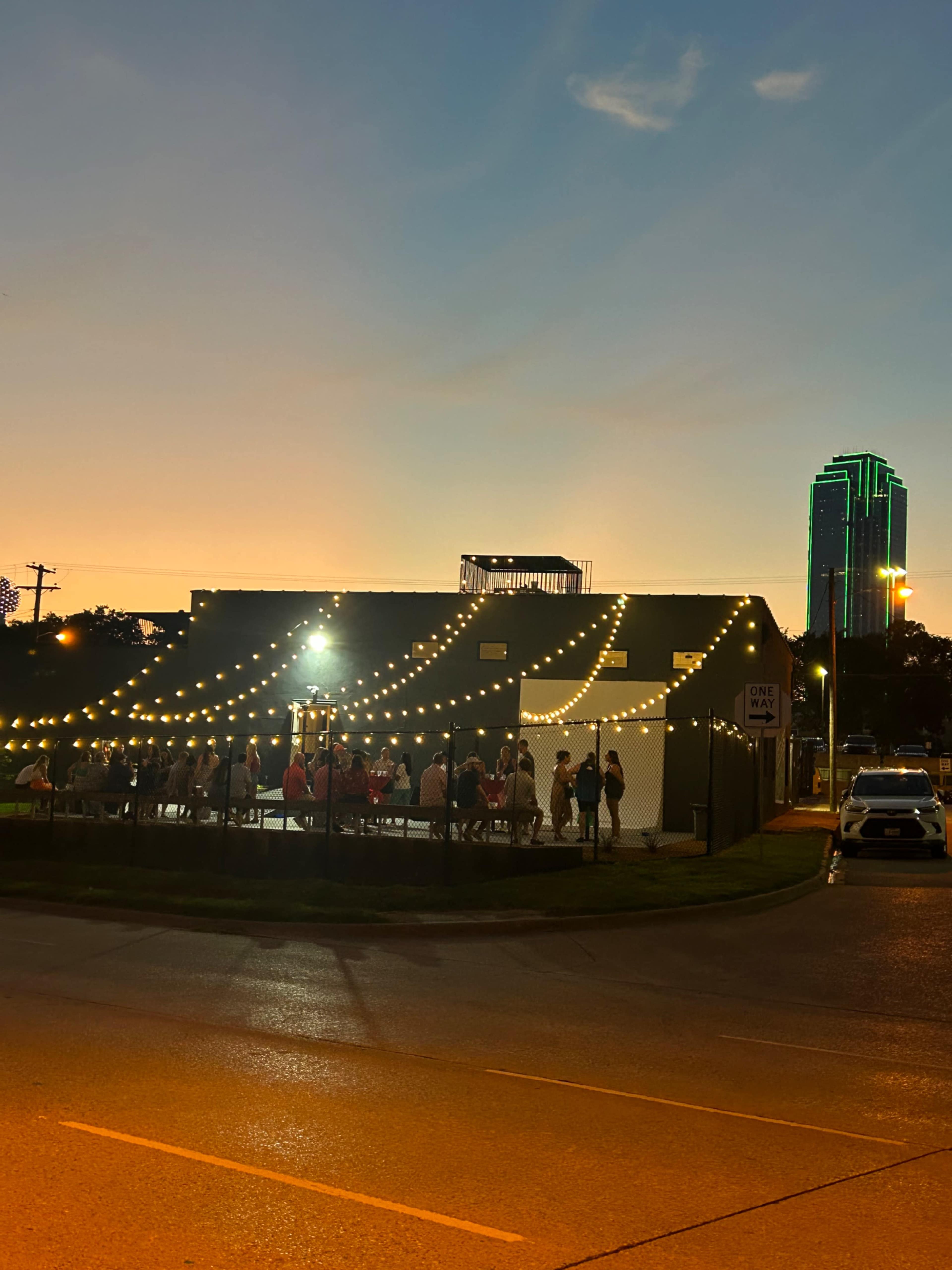 A group of people gathers outdoors under string lights near a building with a skyline featuring green-lit towers at sunset.