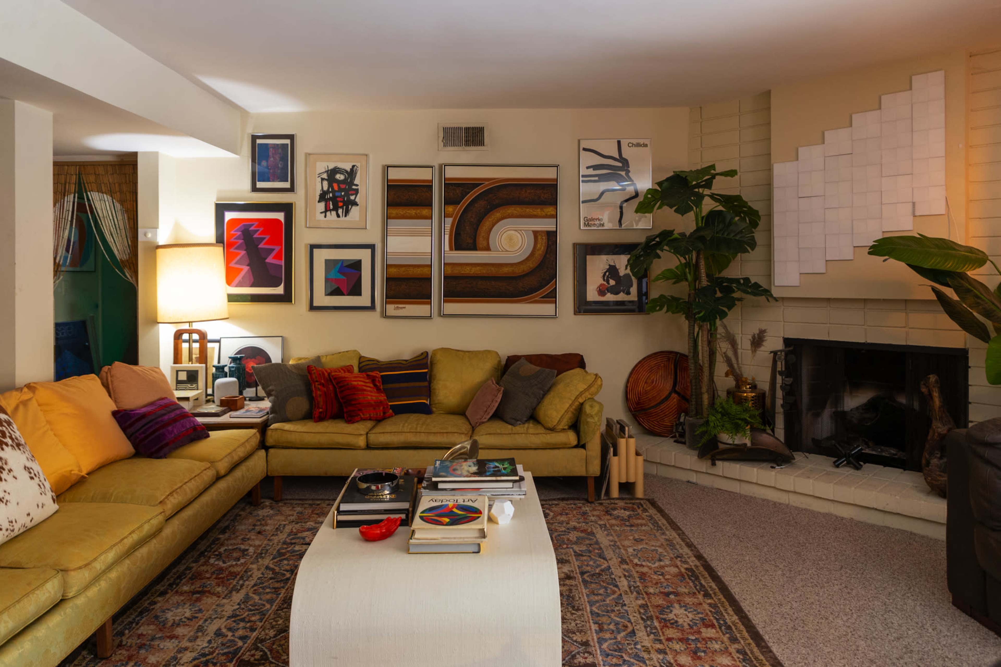 The living room features a green sectional sofa, a coffee table with various books and decor, a fireplace, and an arrangement of colorful artwork on the walls.