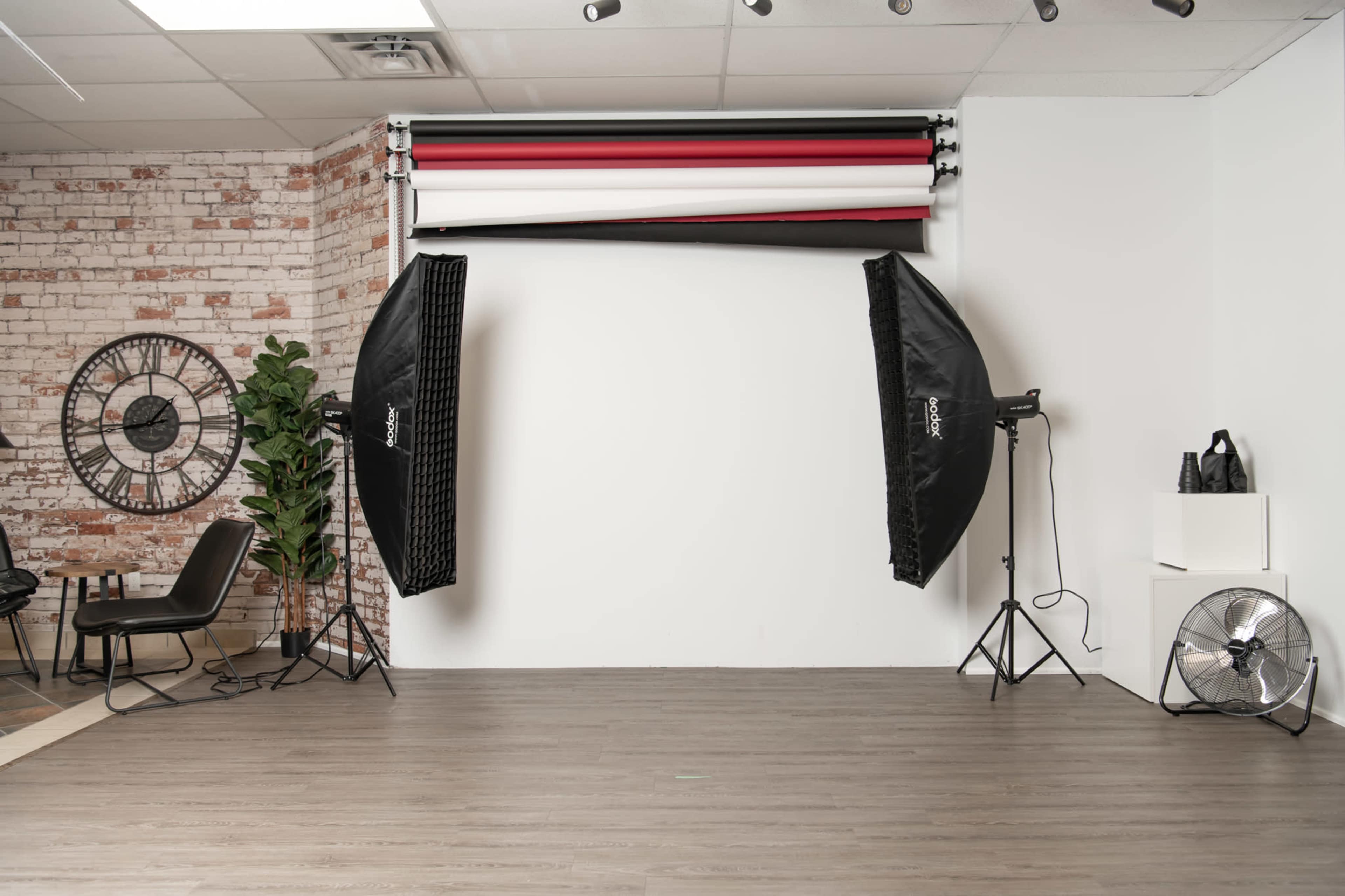 A photography studio featuring a white backdrop, two large softbox lights, a fan, and a few pieces of studio furniture.