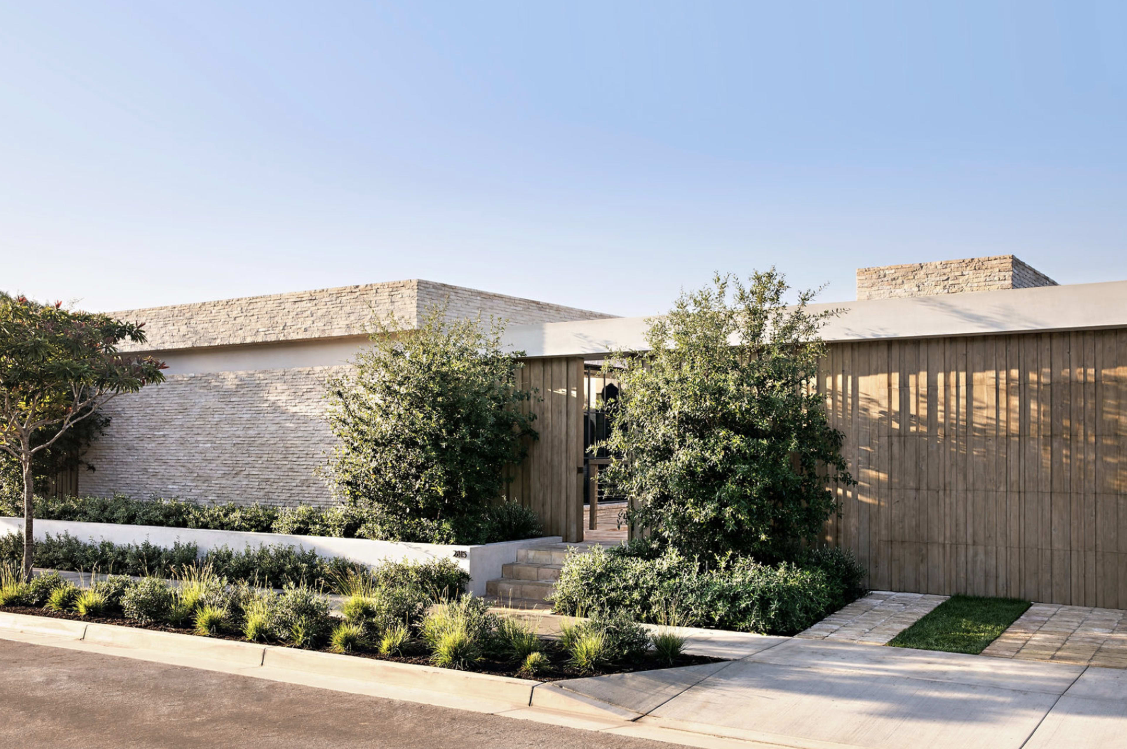 A modern house features a combination of stone and wood facade, surrounded by landscaped greenery and a pathway leading to the entrance.