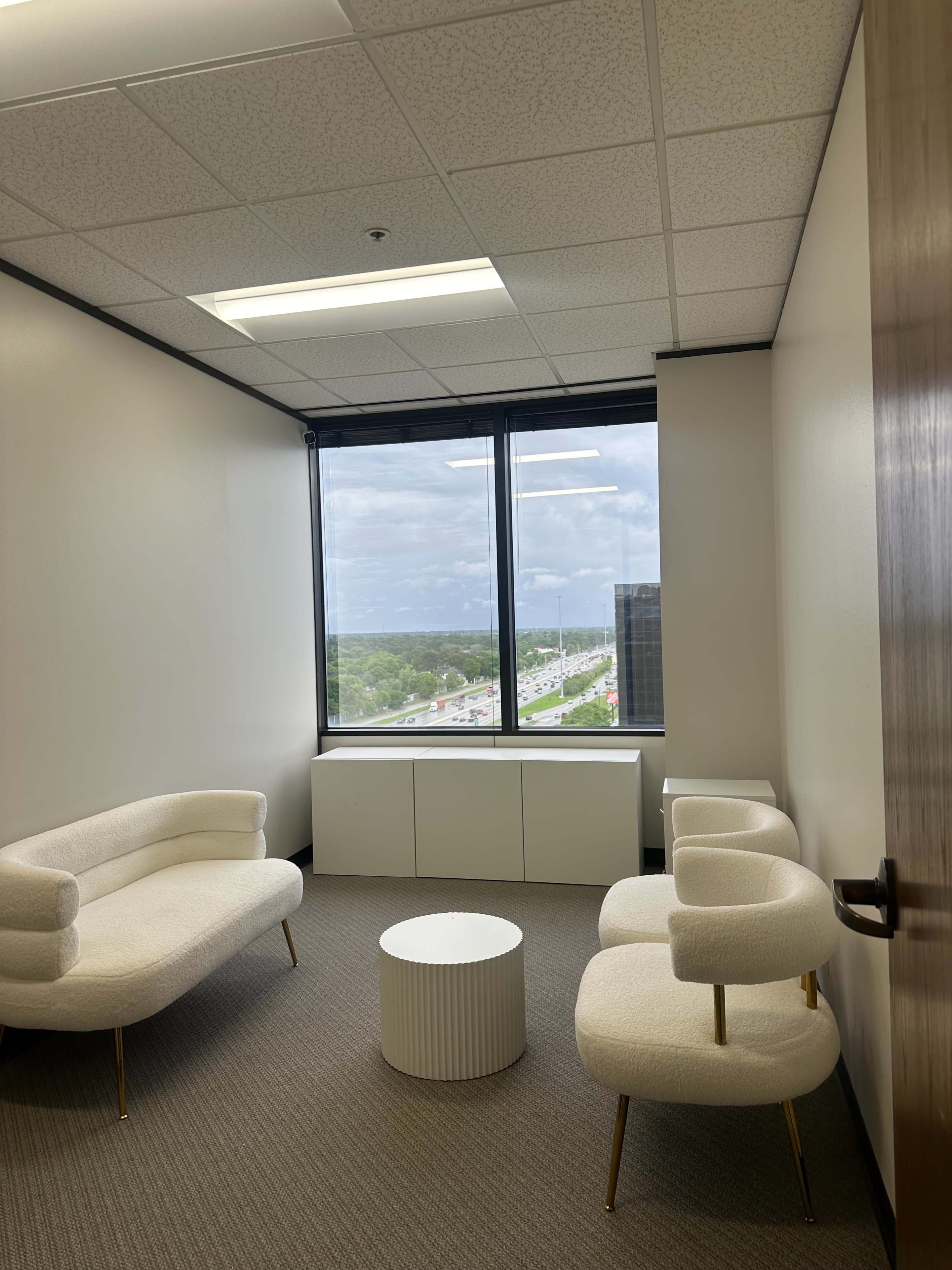 A small office meeting space with two white sofas, two armchairs, and a round table, alongside a window overlooking a highway and green landscape.