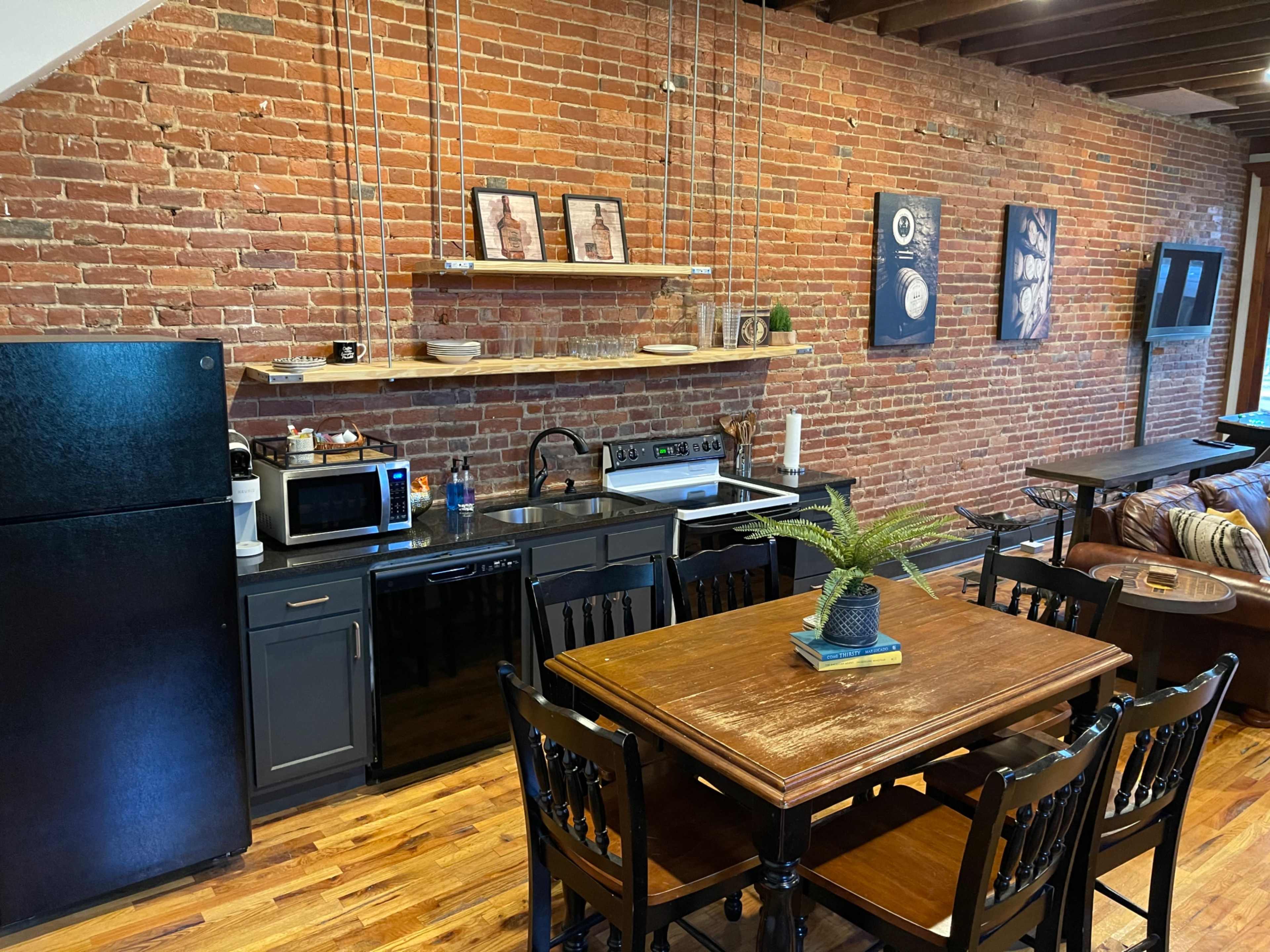A kitchen area with exposed brick walls, a black refrigerator, dark cabinetry, and a wooden dining table set for four.