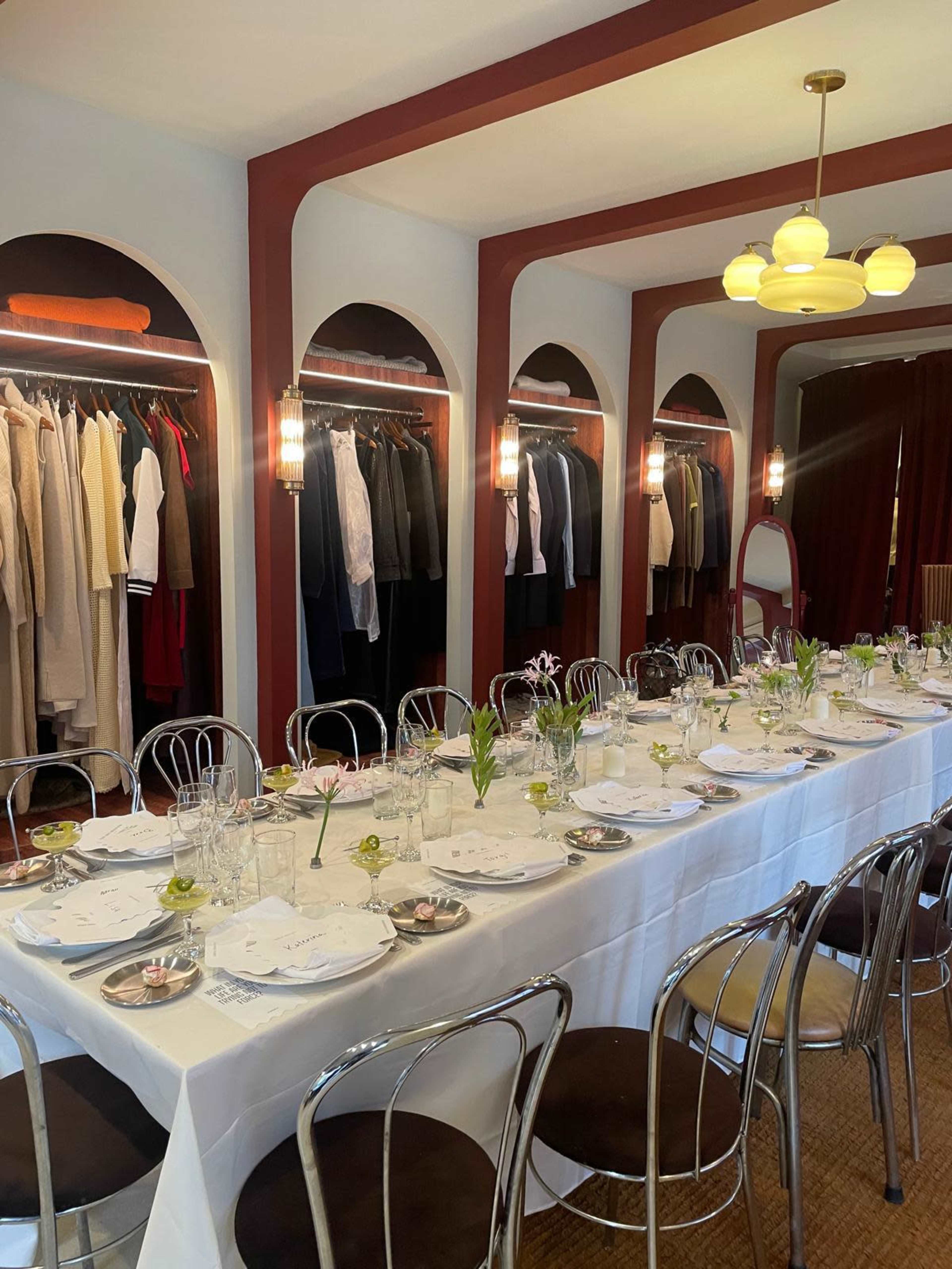 A long dining table is set with plates and glasses, surrounded by a backdrop of neatly arranged clothing hanging on racks.