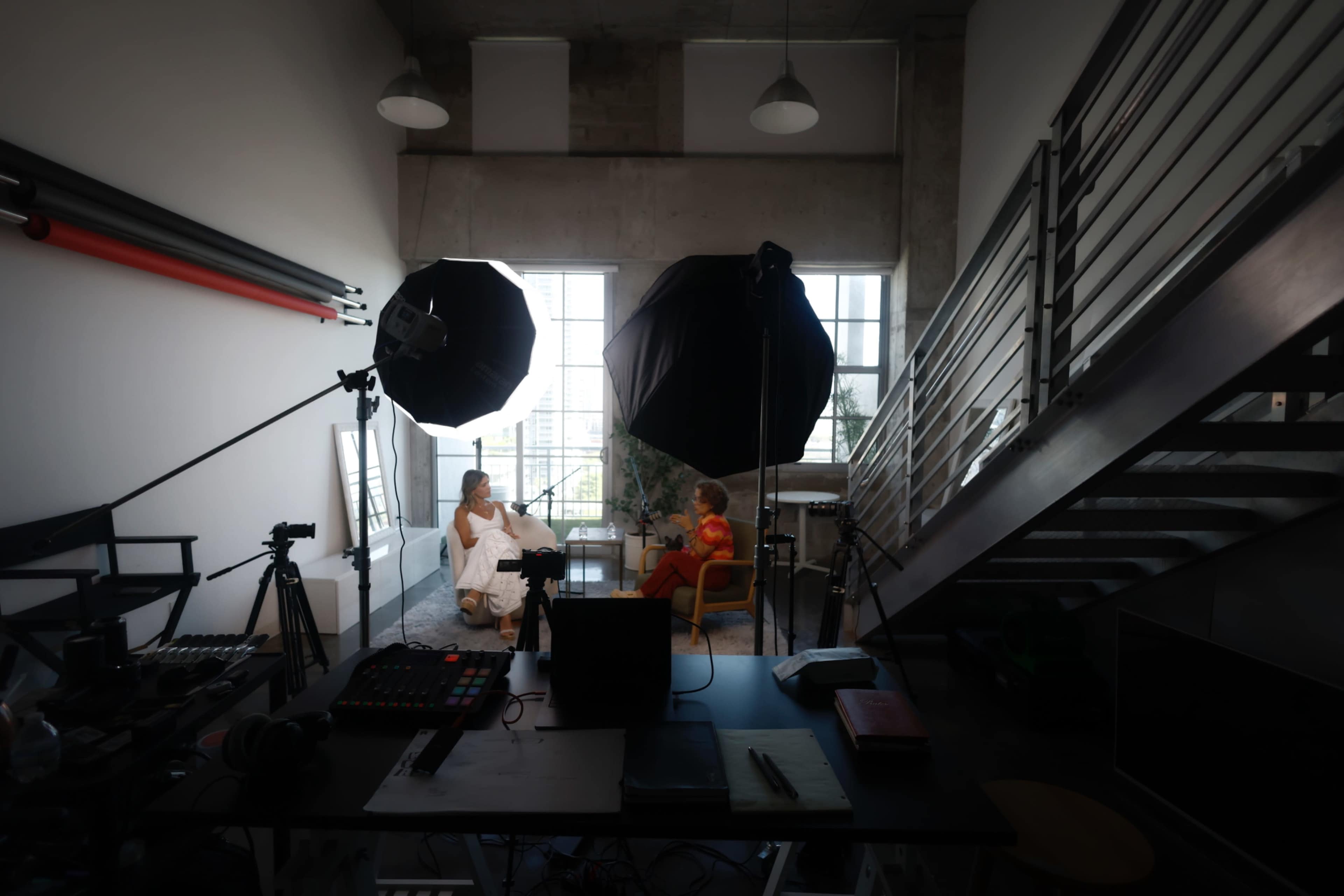 Two women are seated in a brightly lit studio, surrounded by photography equipment and a staircase.