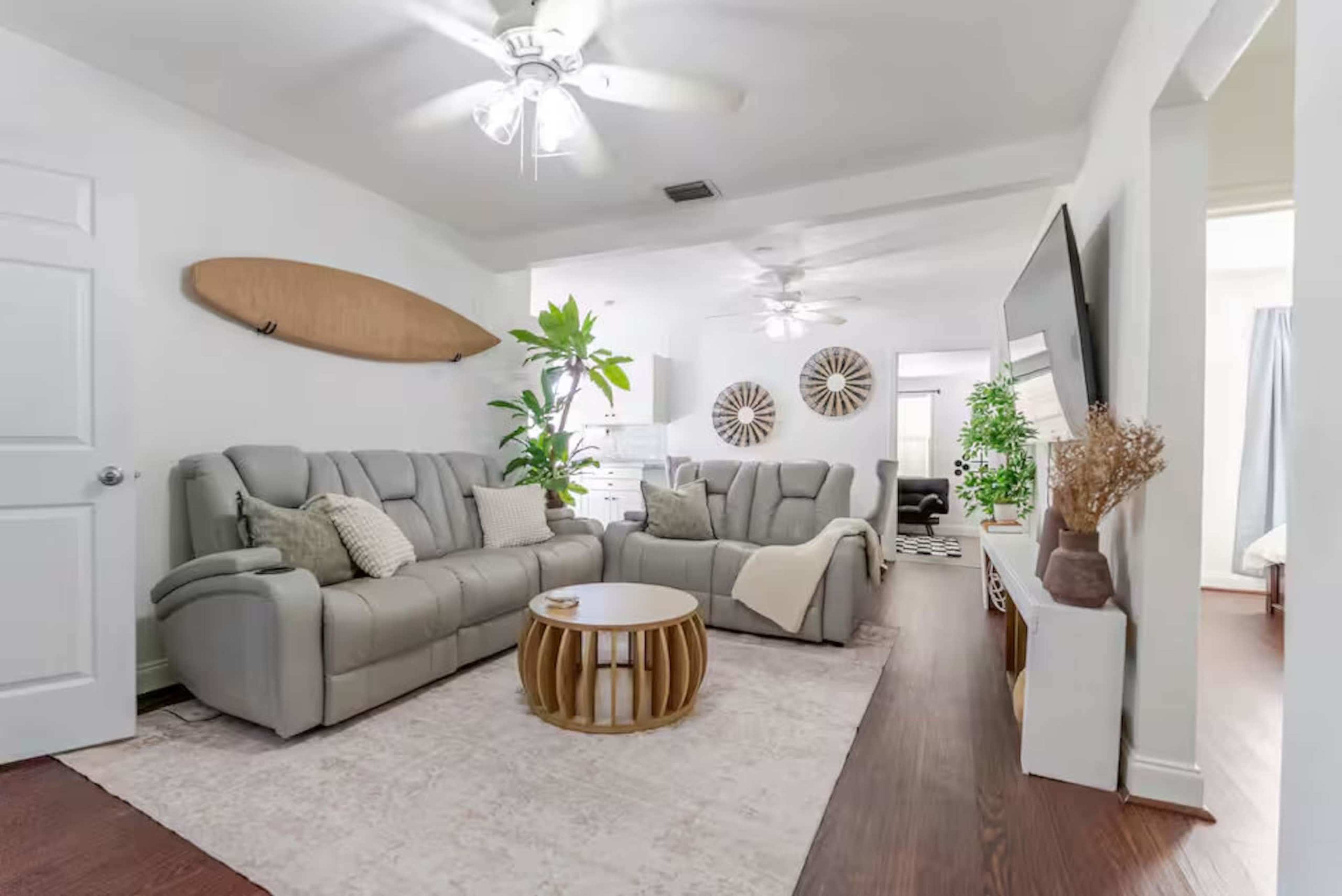 The living room features a light-colored sofa set, a round wooden coffee table, a surfboard on the wall, and decorative plants, with a bright, open layout.