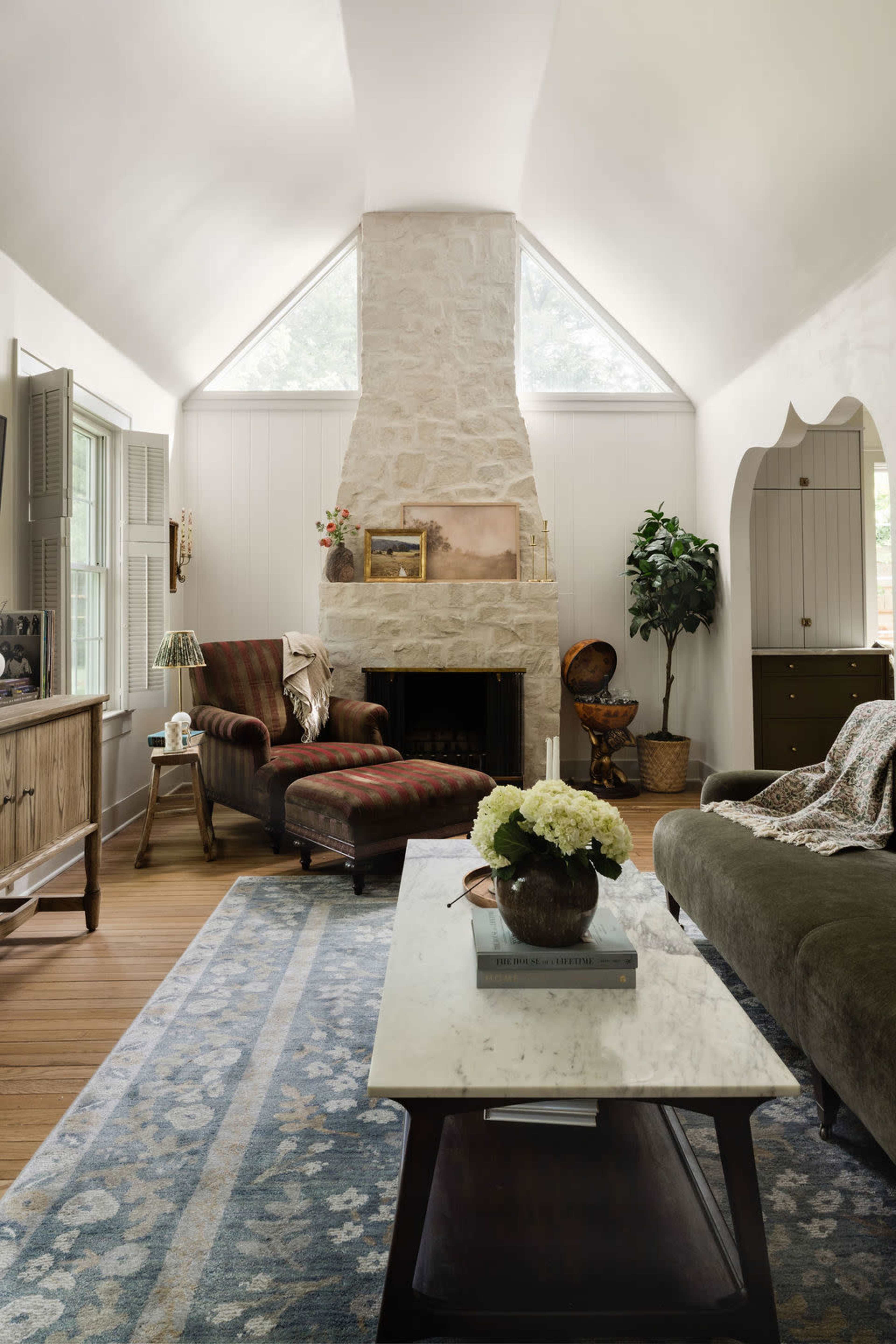 The image depicts a spacious living room featuring a stone fireplace, a mix of cozy seating options, and a coffee table on a patterned rug.