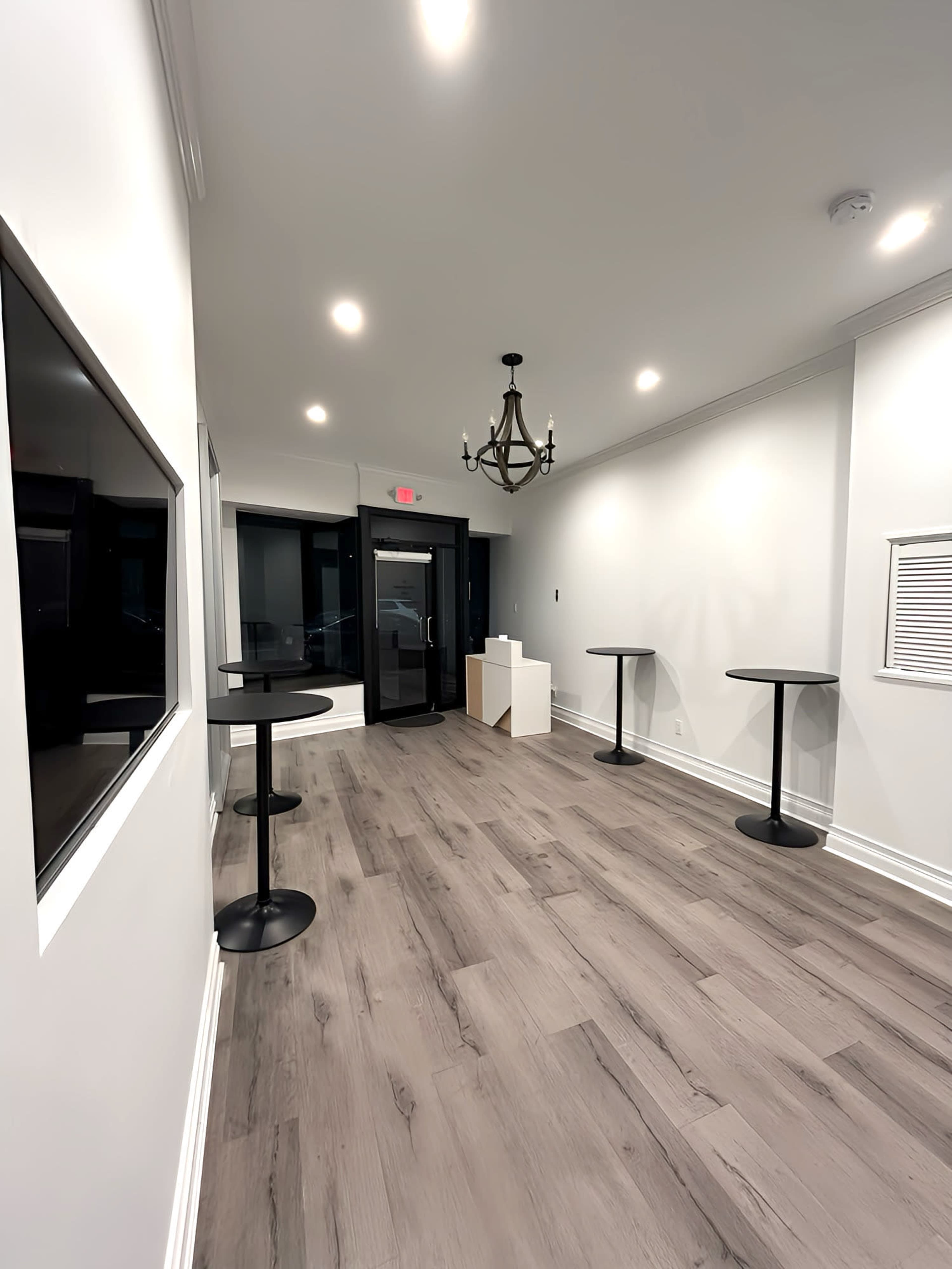 The image shows an empty, well-lit interior hallway with two tall tables, a chandelier, and an entrance door at the far end.