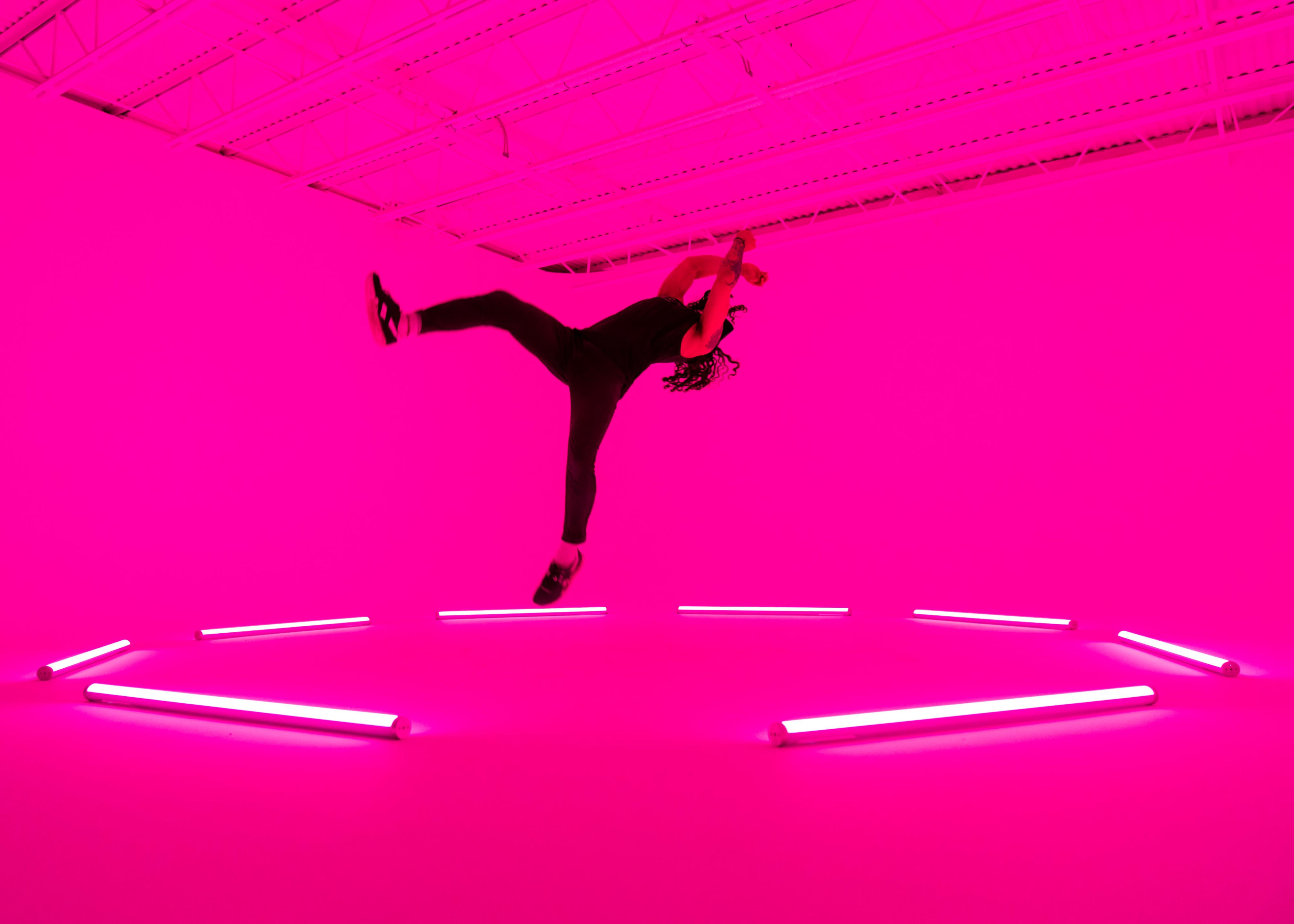 A person performs a dance move in a brightly lit room with pink walls and neon lights.