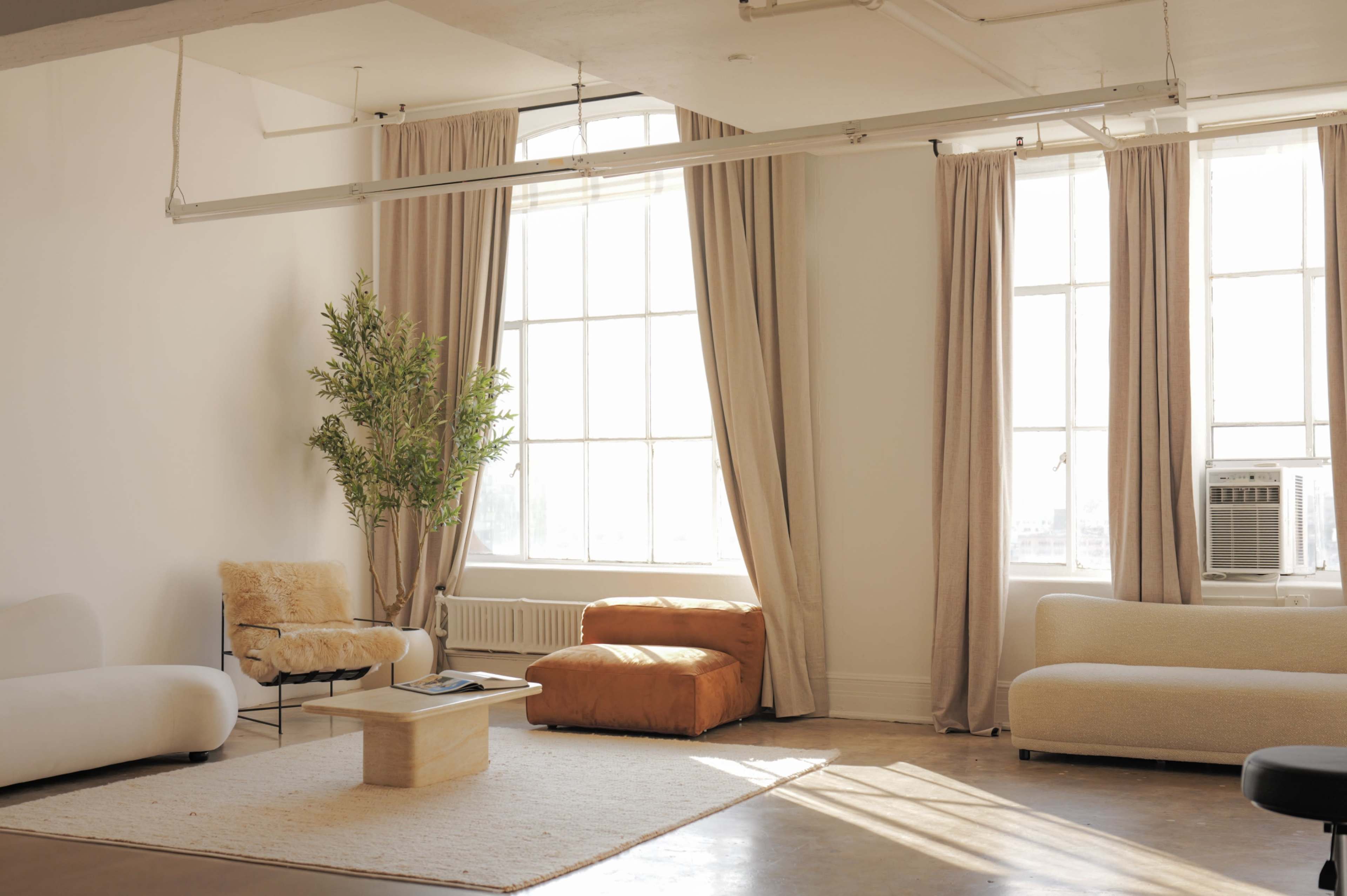 The image shows a minimalist living room with large windows, soft furnishings, and a potted plant.