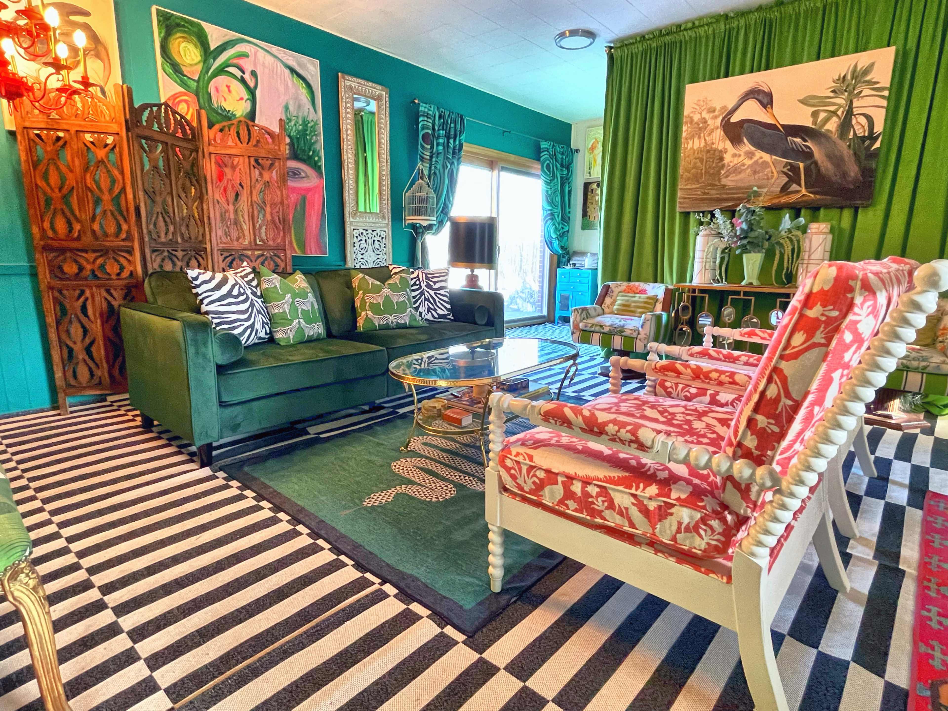 A brightly colored living room featuring a green sofa, striped area rug, and decorative chairs with floral patterns, surrounded by vibrant artwork and greenery.
