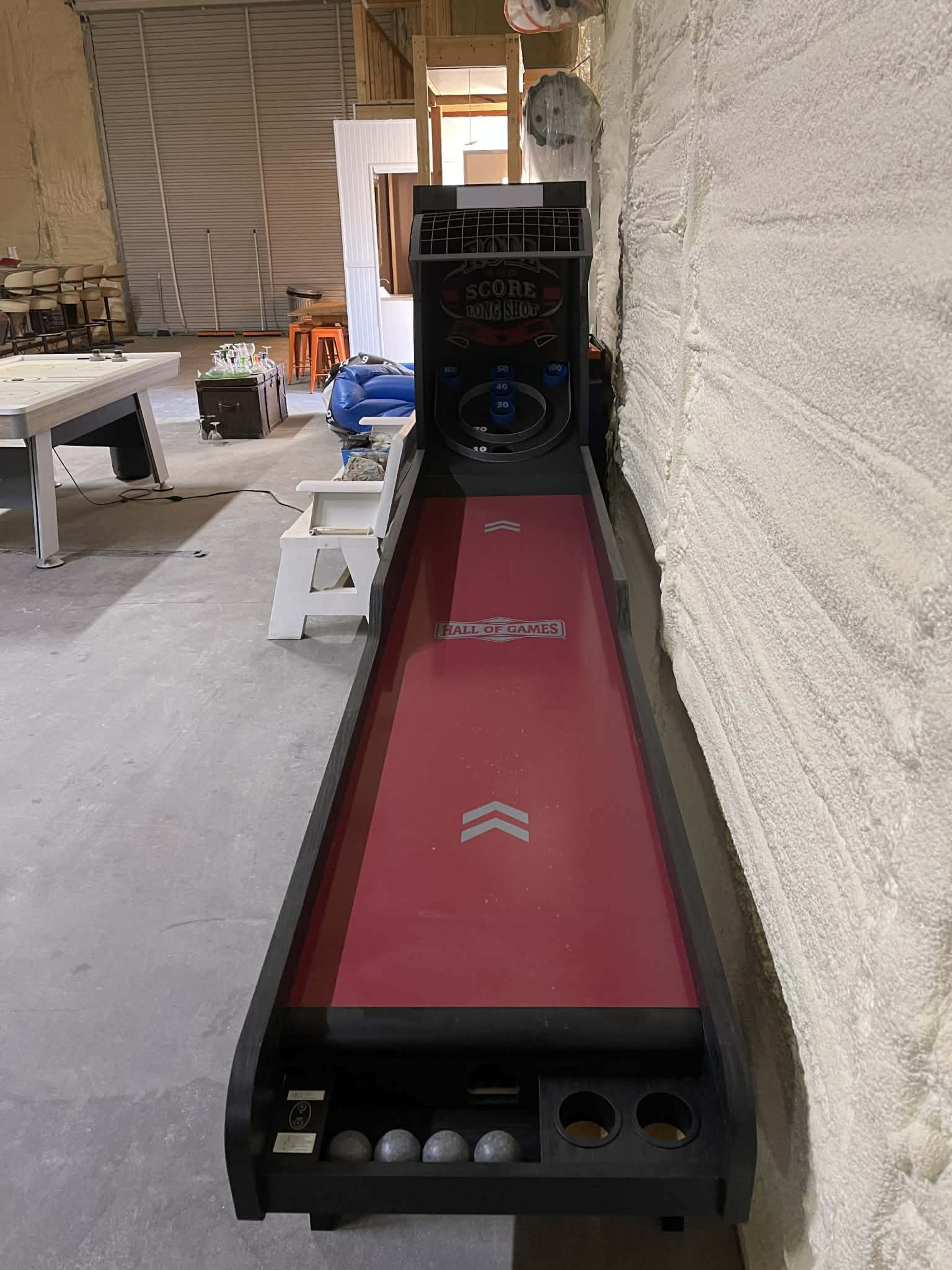 A shuffleboard table is positioned against a wall in a basement or recreational room, with various gaming equipment visible in the background.