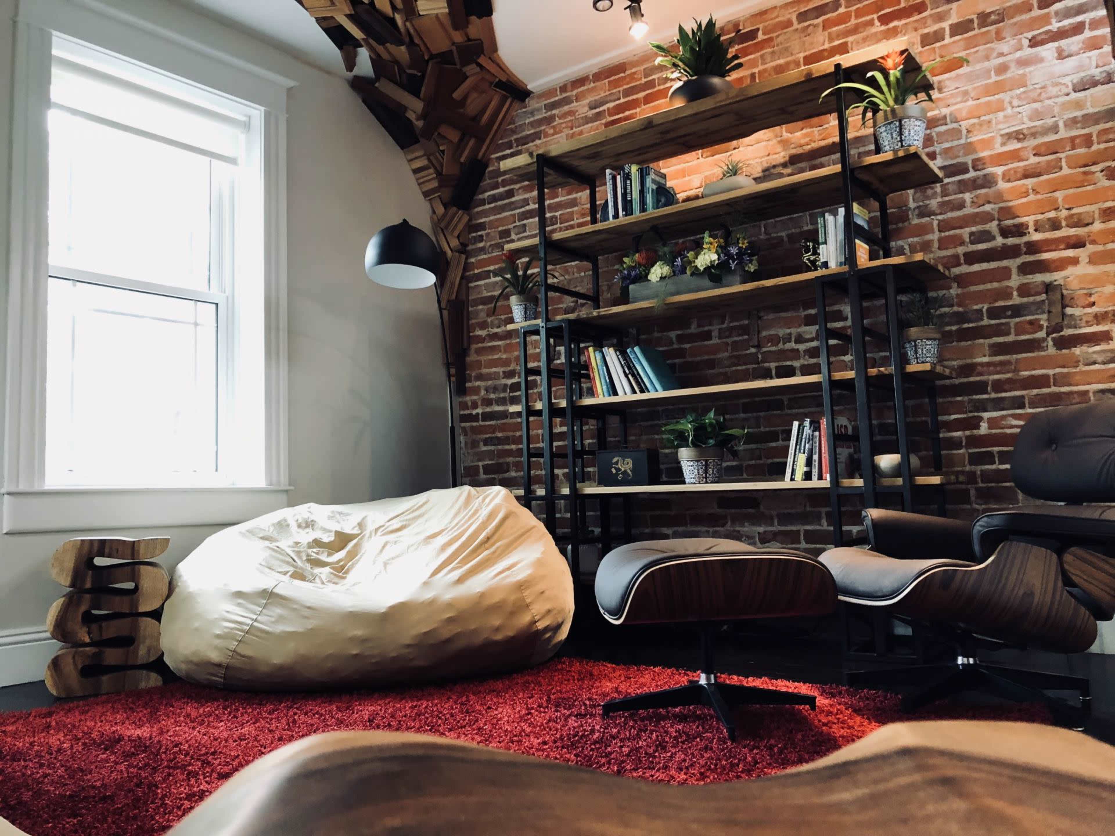The image shows a cozy room with a brick wall, a bookshelf filled with plants and books, a bean bag chair, and a red carpet.