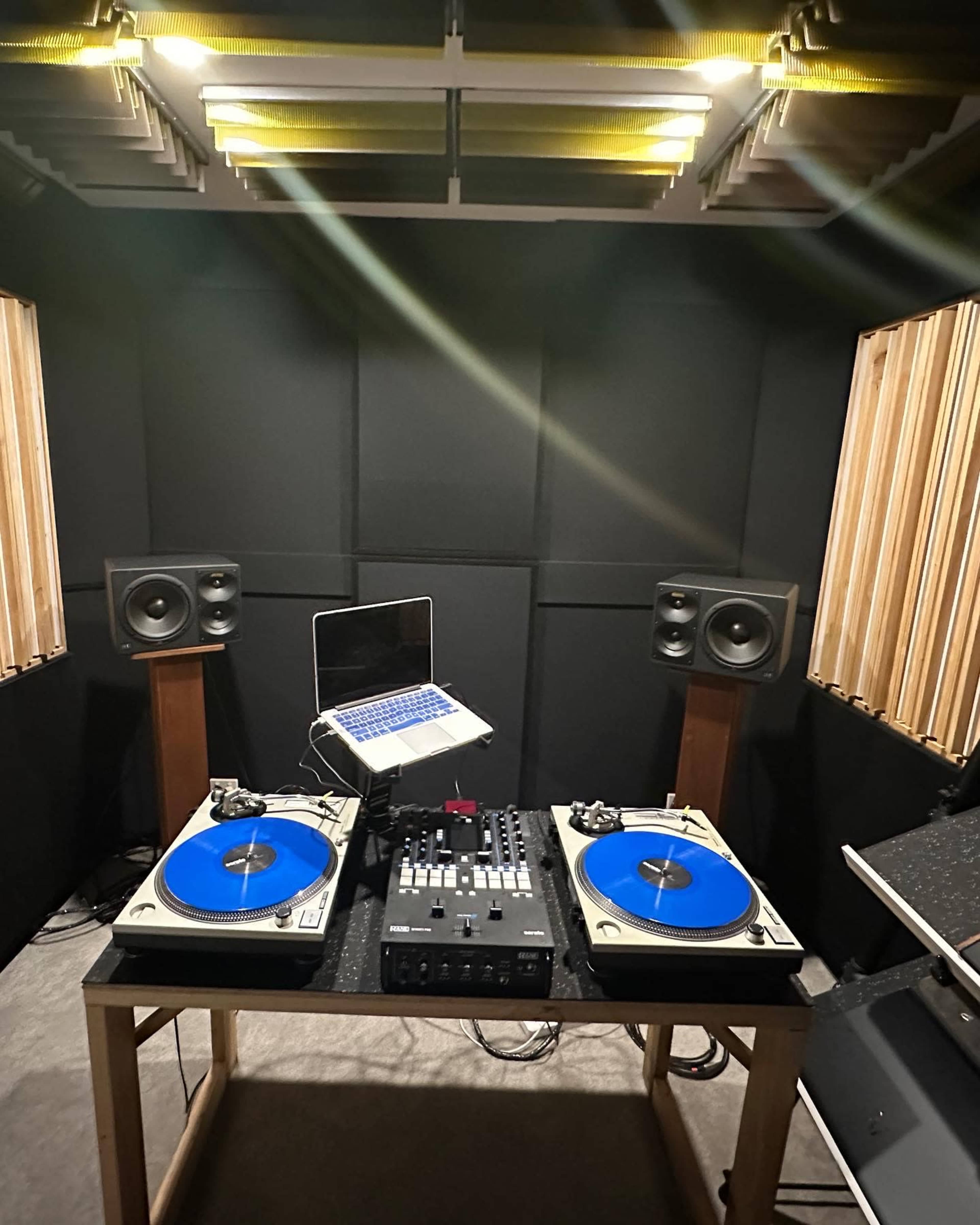 The image shows a DJ setup featuring two turntables with blue platters, a control mixer, and a laptop, all arranged in a soundproof room with wooden paneling.
