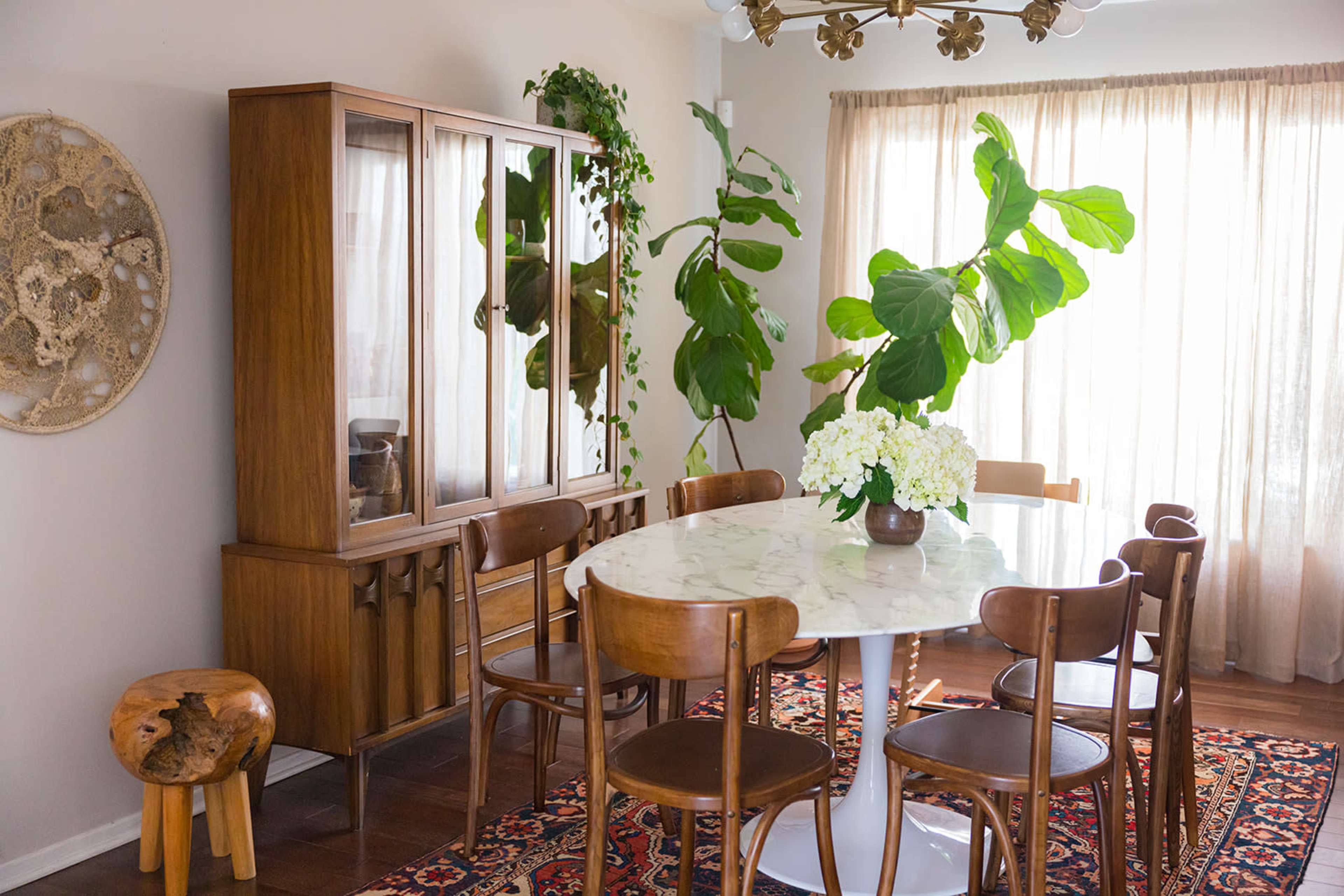 A dining room features a round marble table surrounded by wooden chairs, a glass-fronted cabinet, and large plants near a window with sheer curtains.