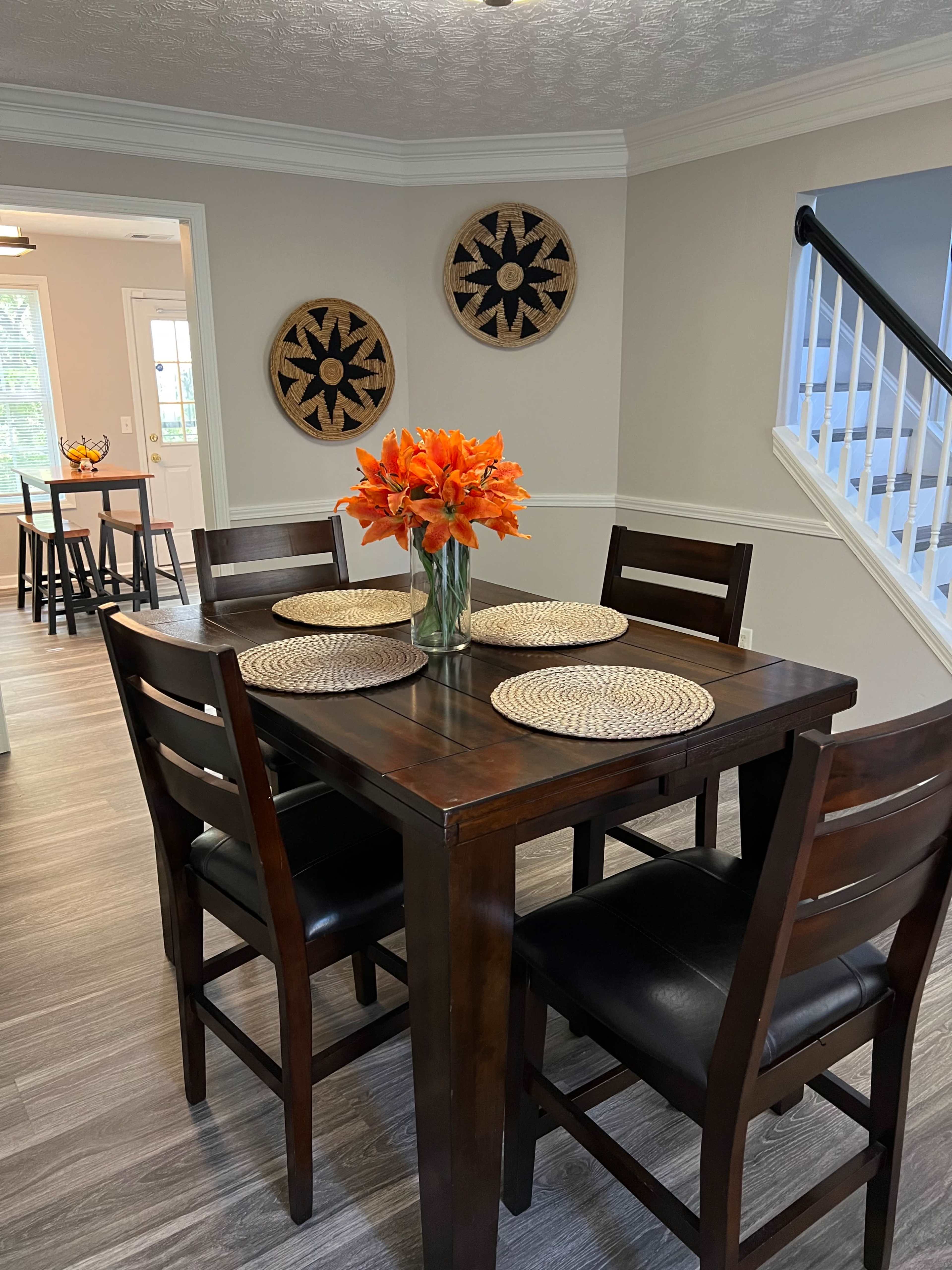 The image shows a dining area featuring a wooden table with four chairs, a vase of orange flowers in the center, and woven placemats on the table.