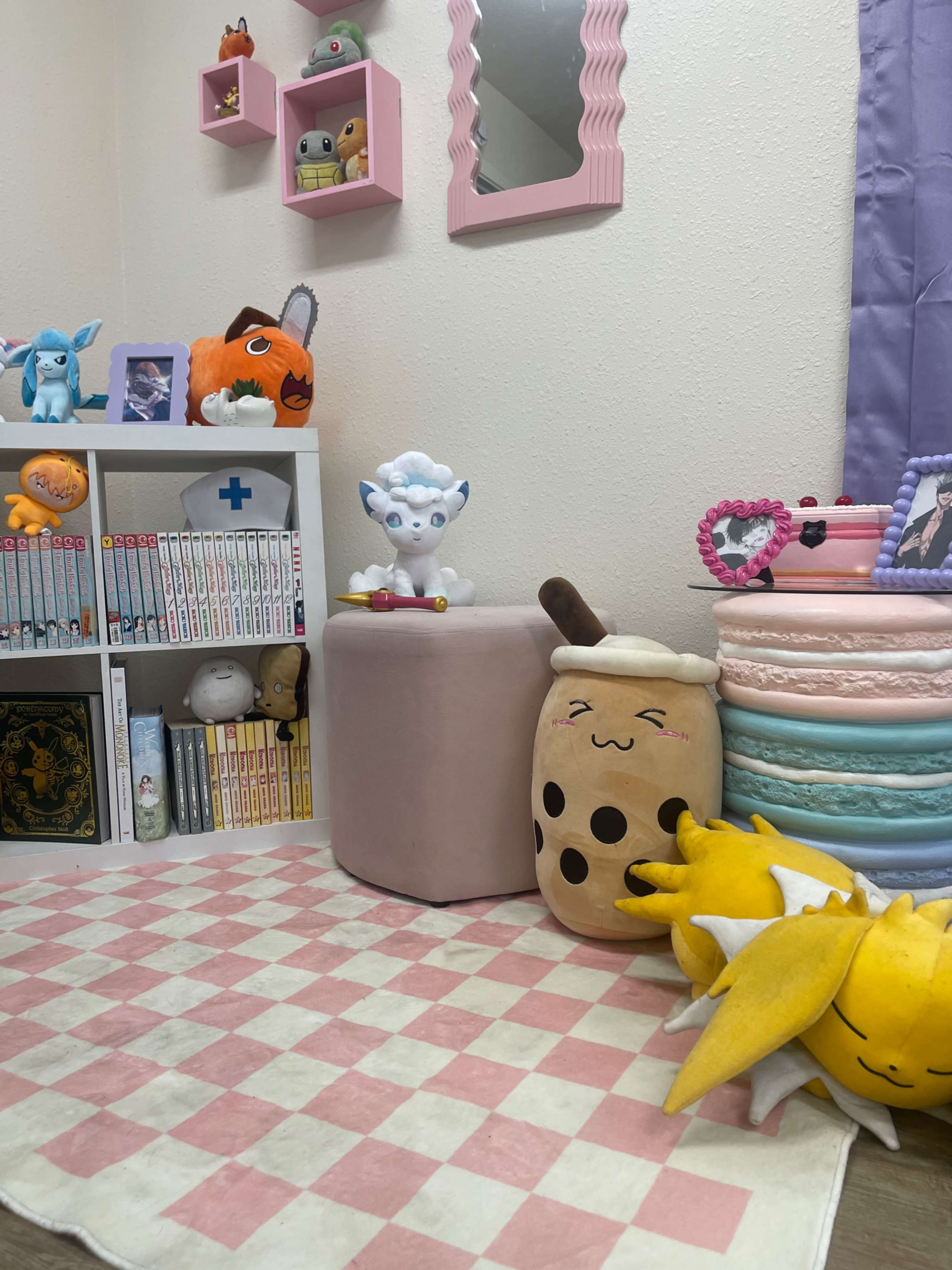 The image shows a colorful room corner decorated with plush toys, a small bookshelf filled with manga, and a pink ottoman next to a stack of pastel-colored macarons.