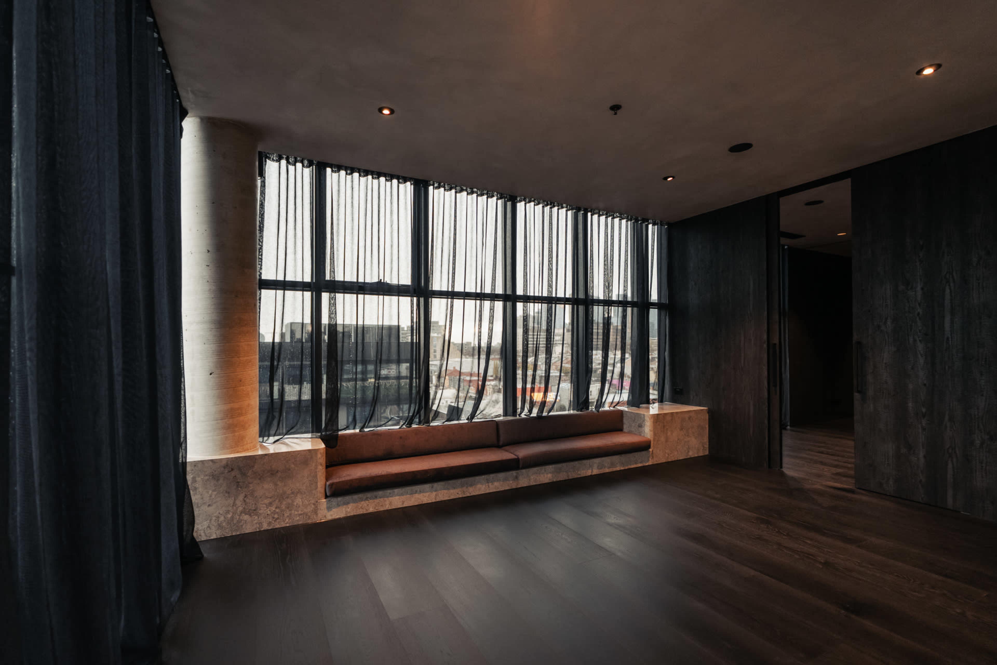 A modern room features a long window with sheer curtains, a concrete bench, and dark wooden flooring.