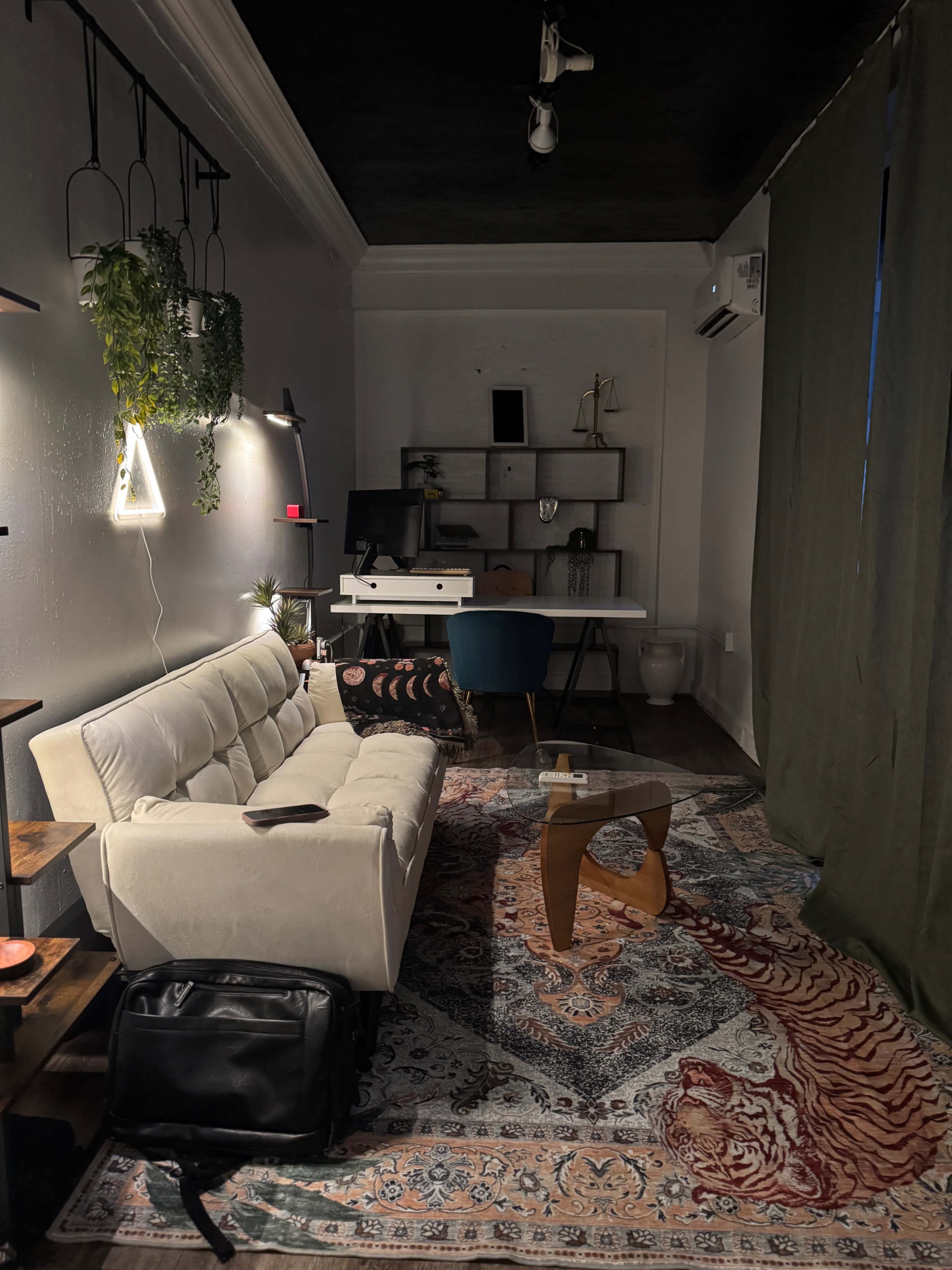 The image shows a small, stylish workspace featuring a white couch, a desk with a computer, and decorative elements, all set against a patterned rug and a green curtain.