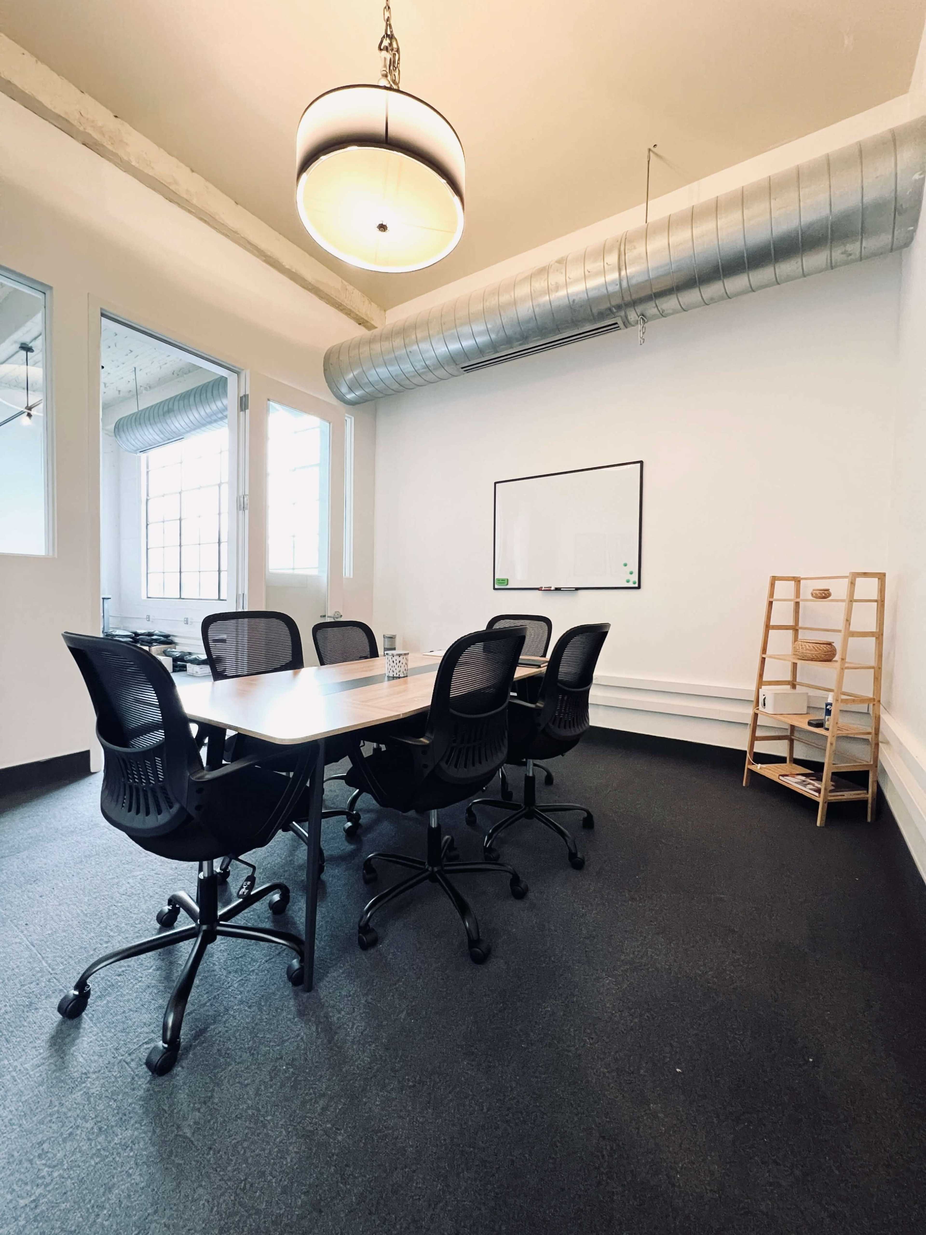 A conference room with a large table surrounded by six black office chairs, a whiteboard on the wall, and a shelving unit in the corner.