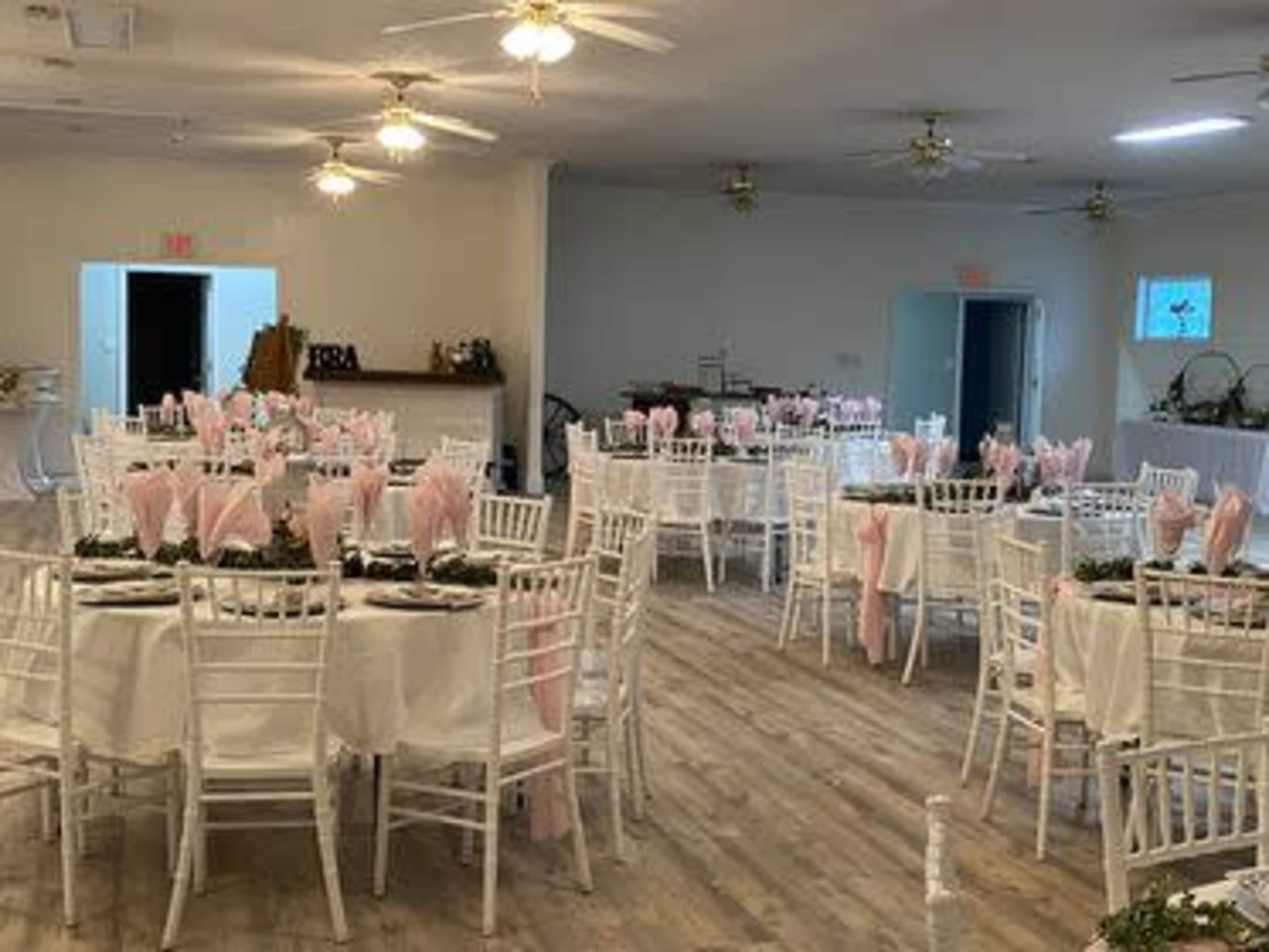 A banquet hall is set up with several round tables, each adorned with white tablecloths, pink napkins, and greenery.