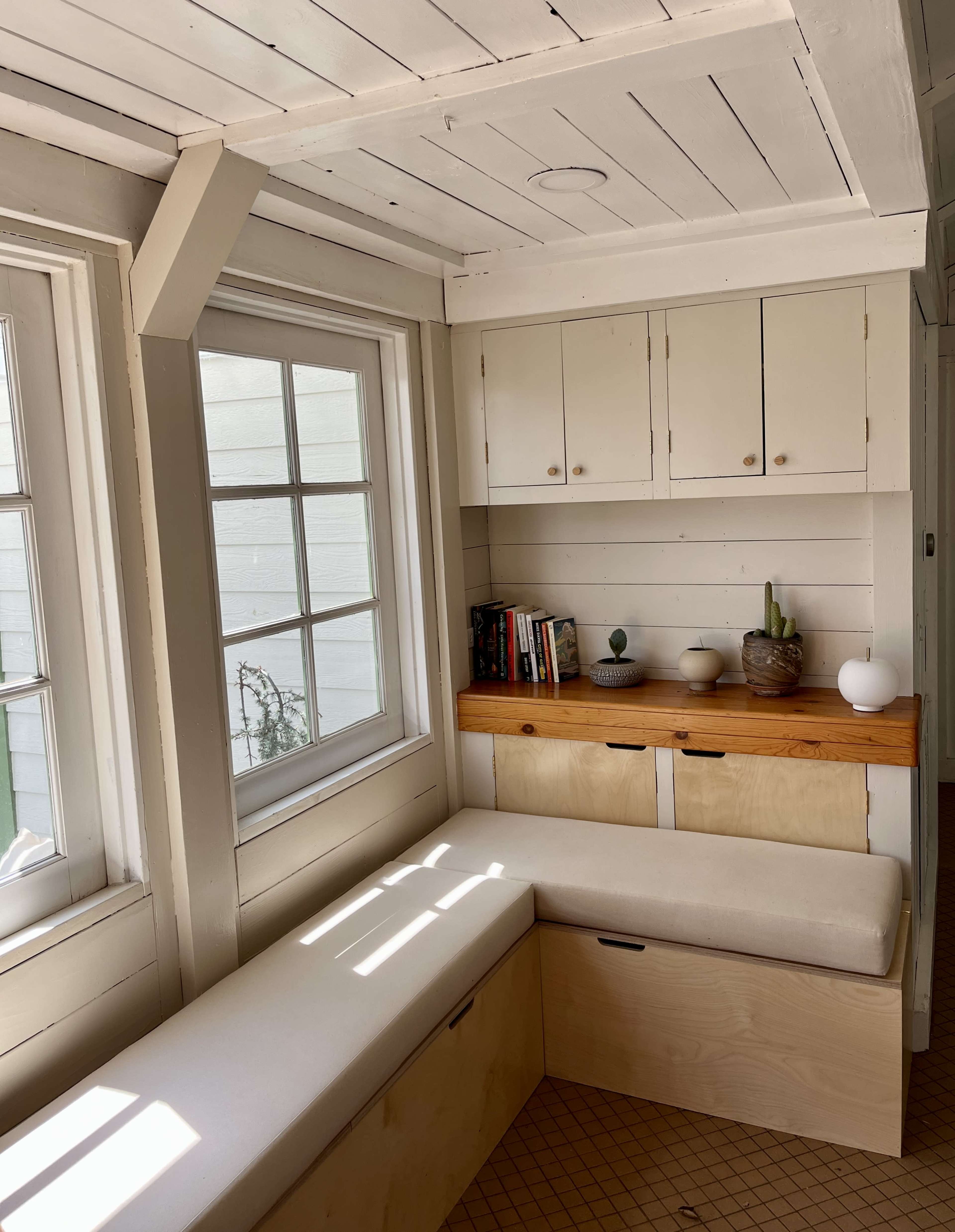 A bright, minimalist corner with a wooden bench, a shelf containing books, and several decorative items against a backdrop of white walls and large windows.