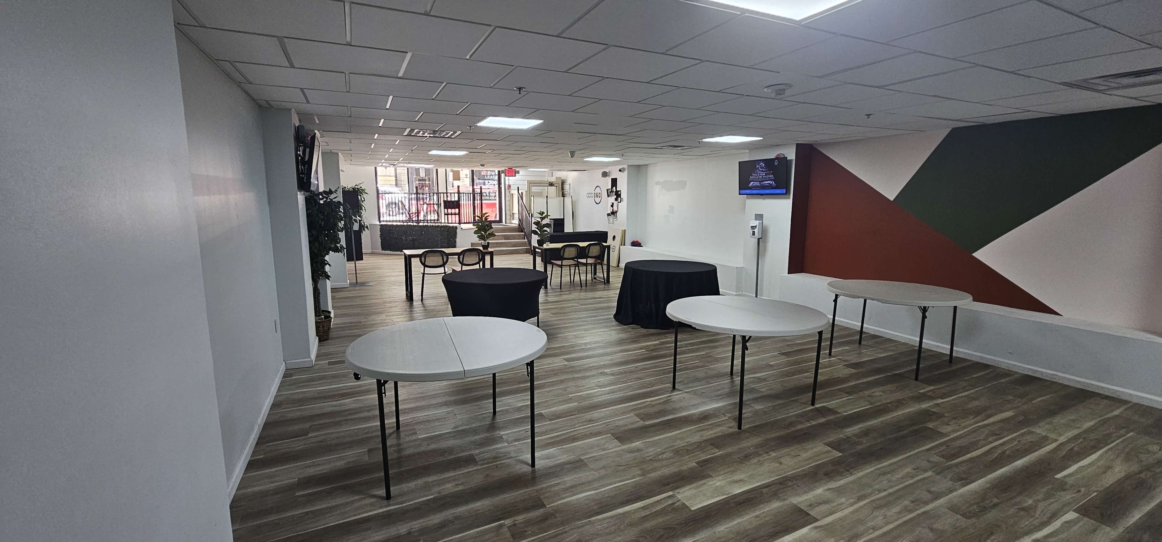 The image shows a spacious interior with several round and rectangular tables arranged, a seating area in the background, and a partially painted wall featuring geometric designs.