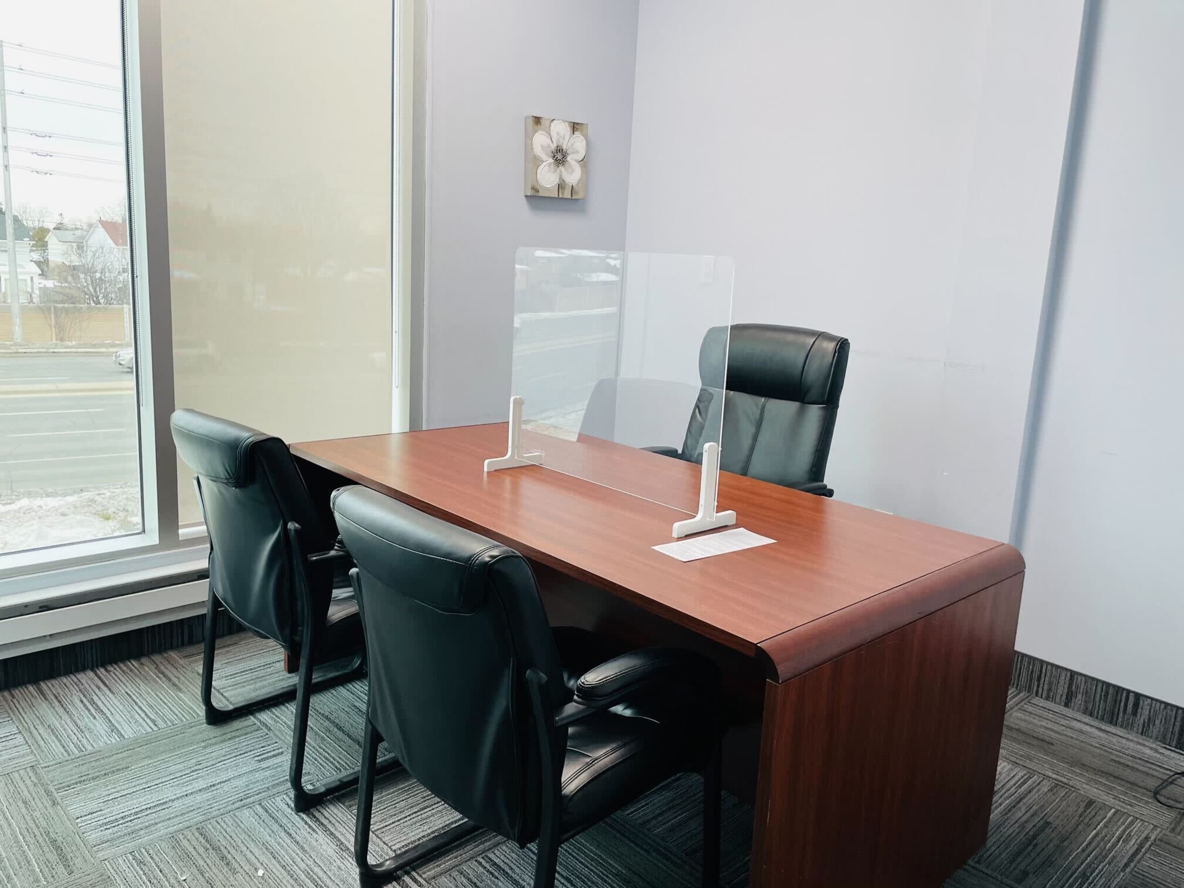 A modern office space features a wooden desk with a protective acrylic screen and two black chairs positioned around it.