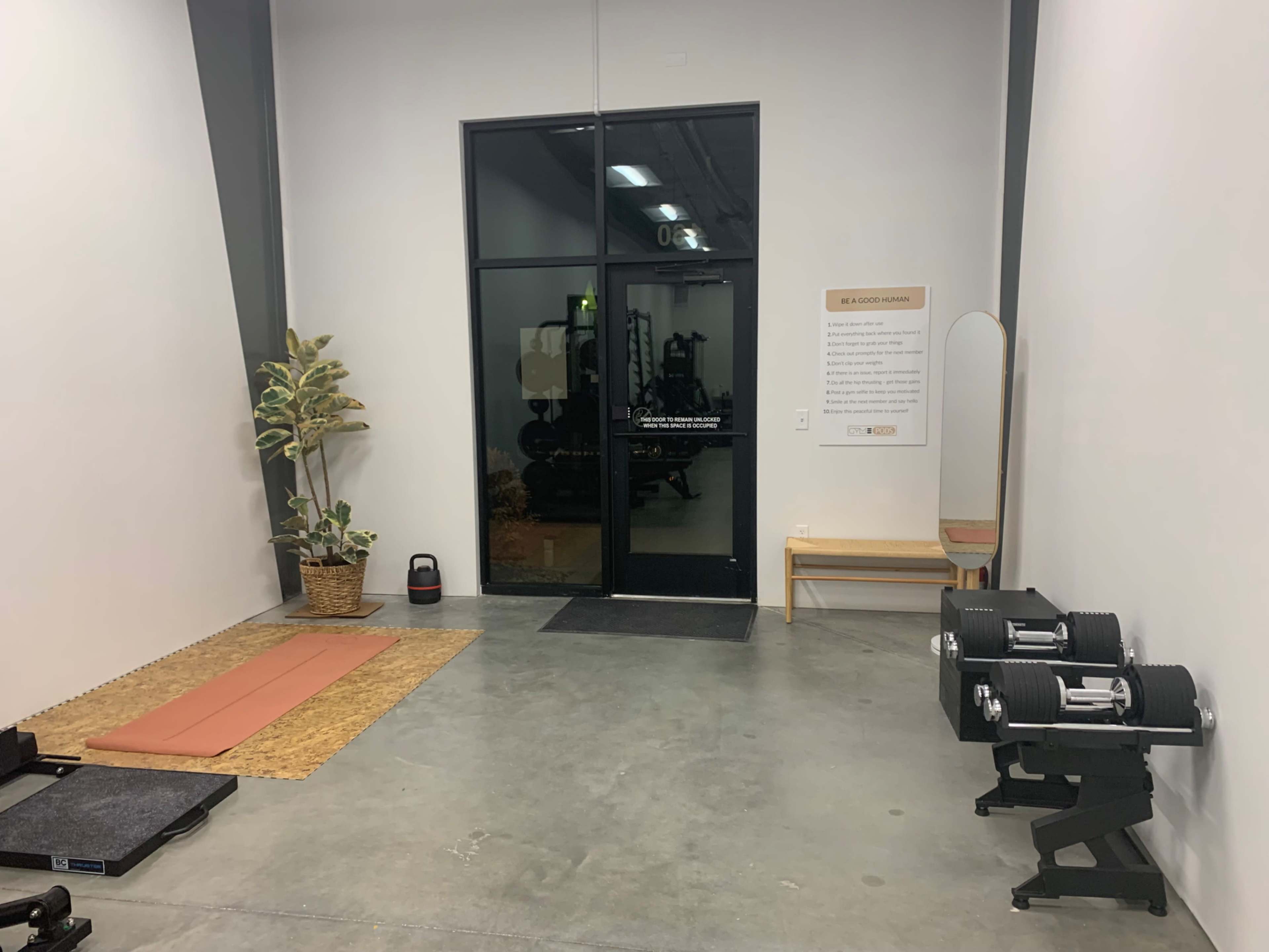 The image shows a minimalistic gym area featuring a yoga mat, dumbbells, a potted plant, a mirror, and a glass door leading outside.