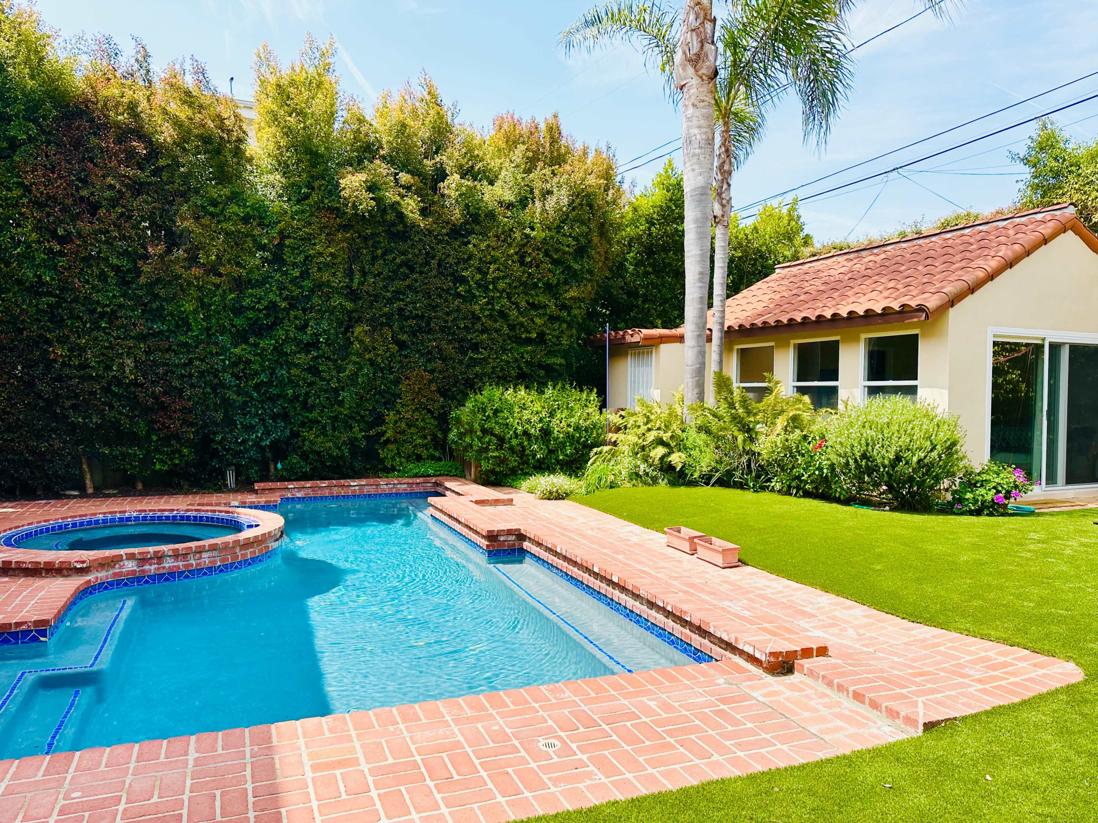 A residential backyard with a swimming pool, spa, and a small house surrounded by greenery.