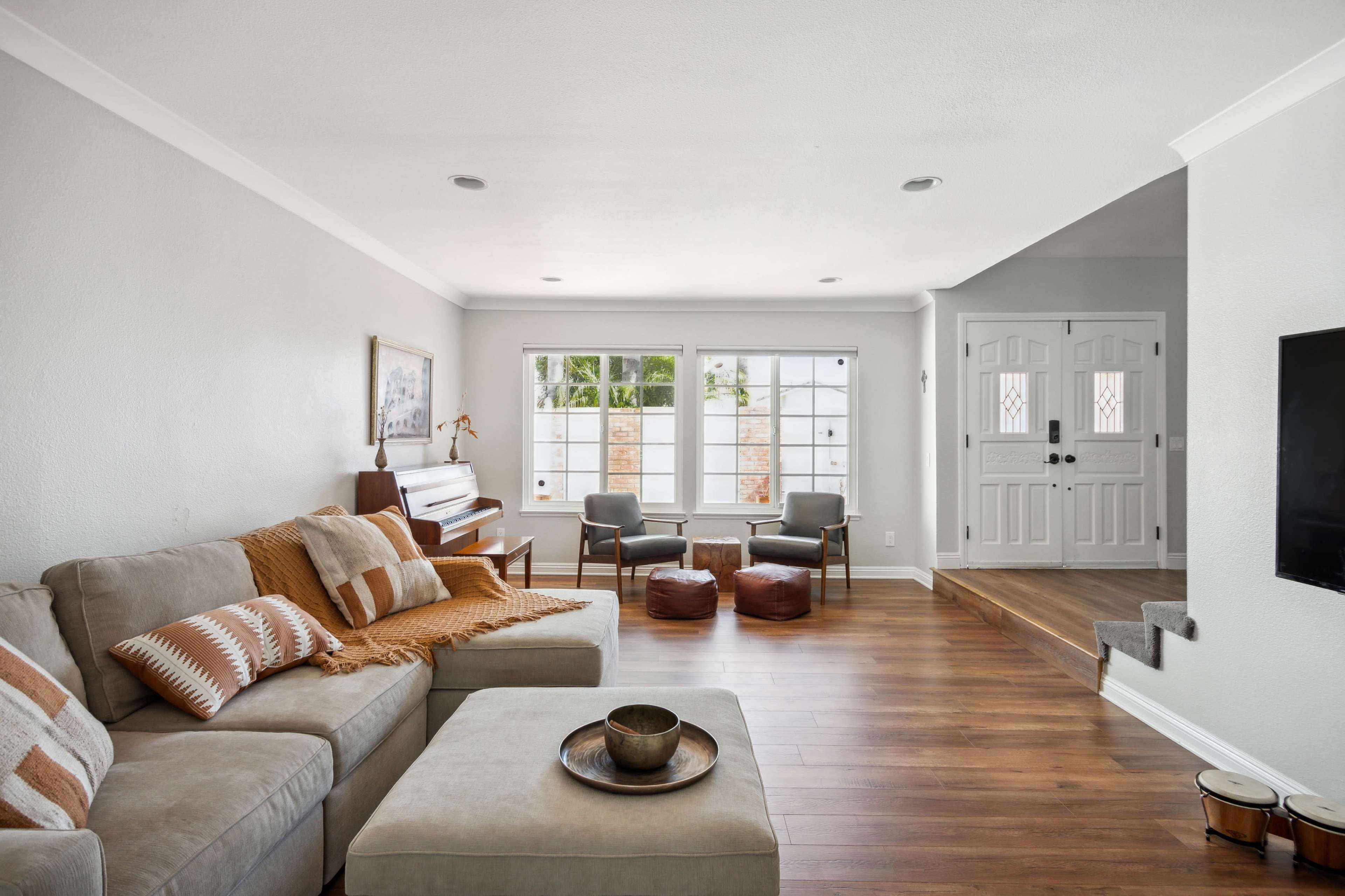 A light-filled living room features a sectional sofa, a piano, and two chairs with a coffee table in front of them.