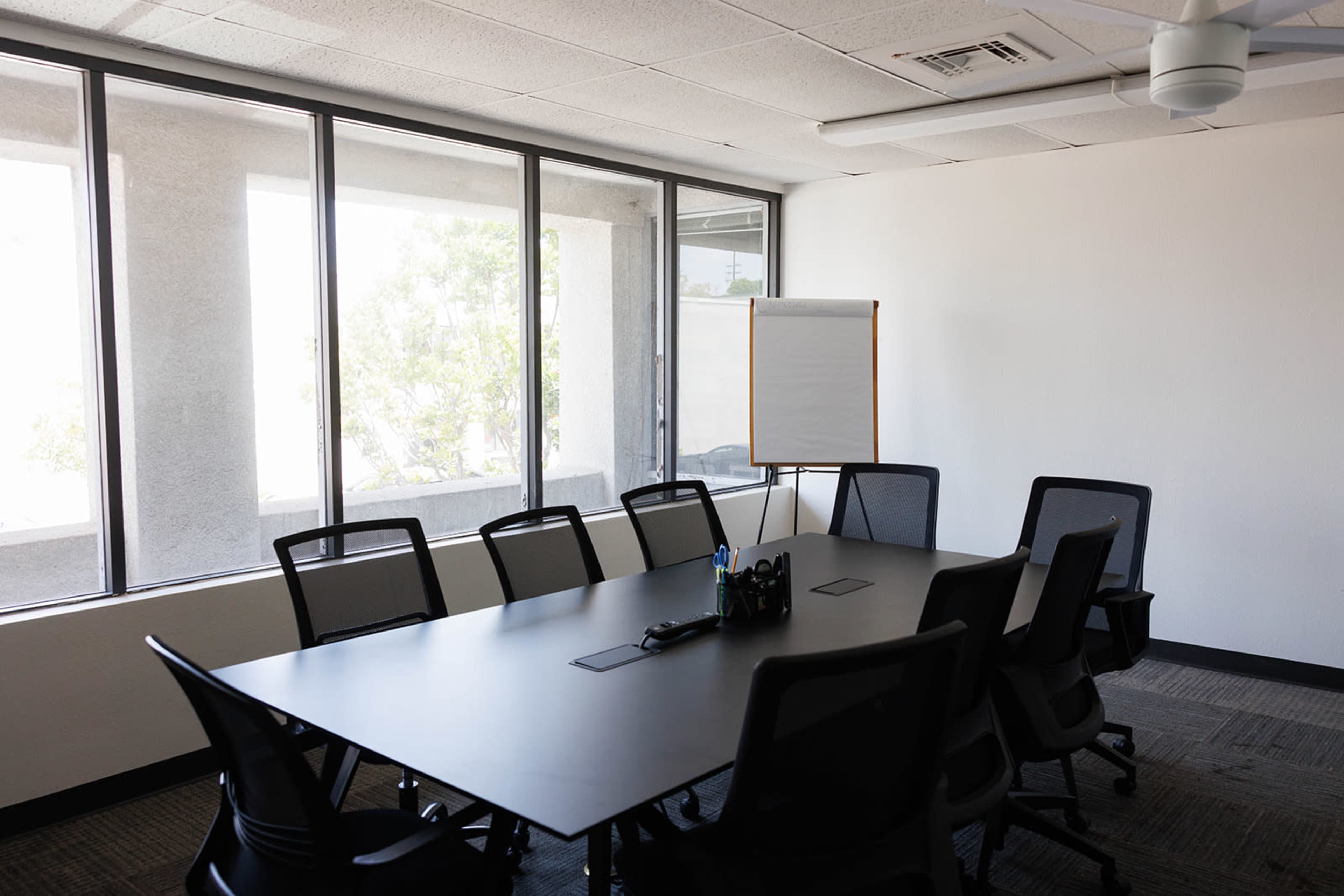 A modern conference room features a large table surrounded by black chairs, with a whiteboard nearby and windows letting in natural light.