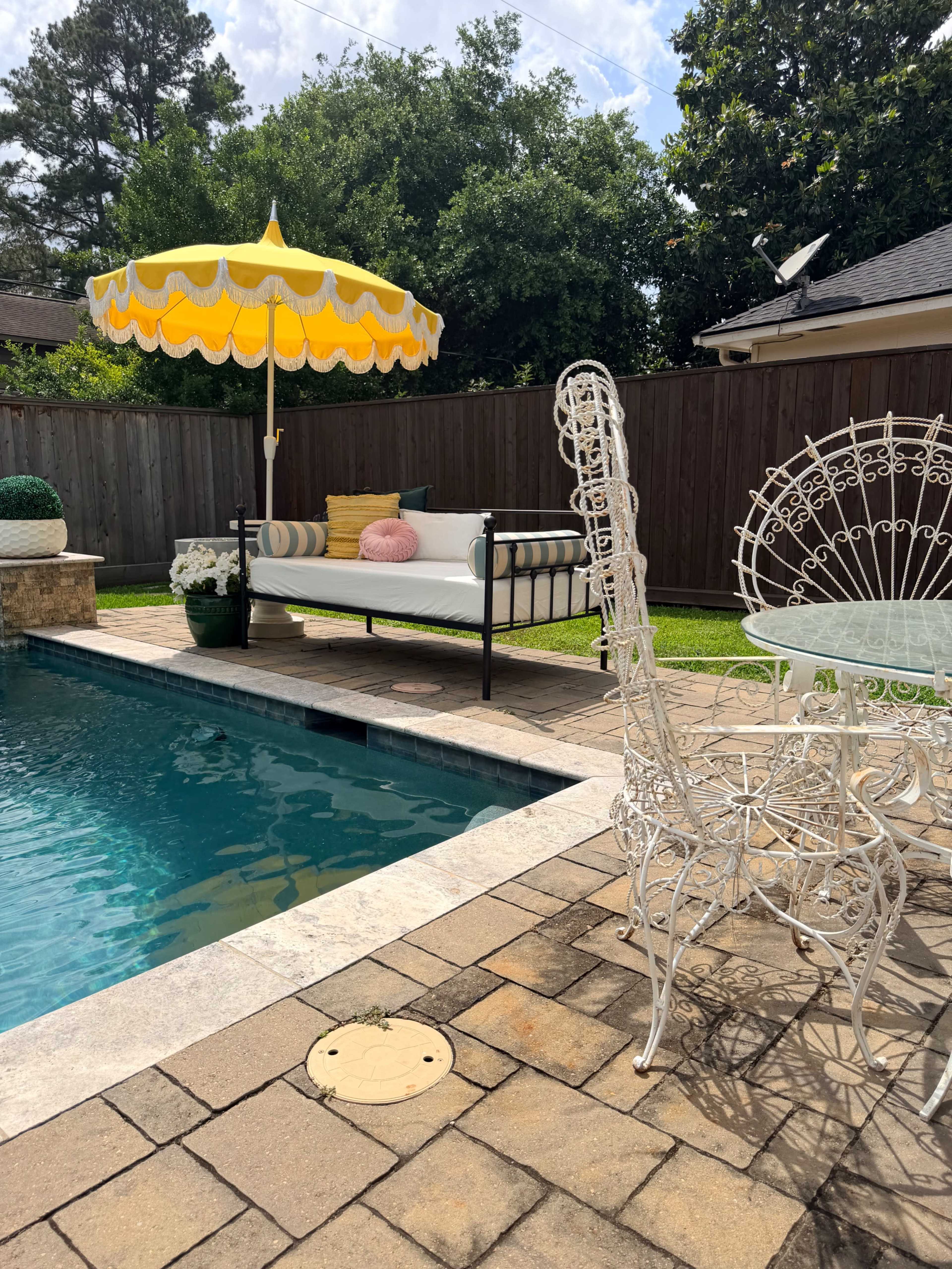 A poolside seating area features a daybed with pillows, an umbrella for shade, and a white wrought iron table and chairs surrounded by lush greenery.