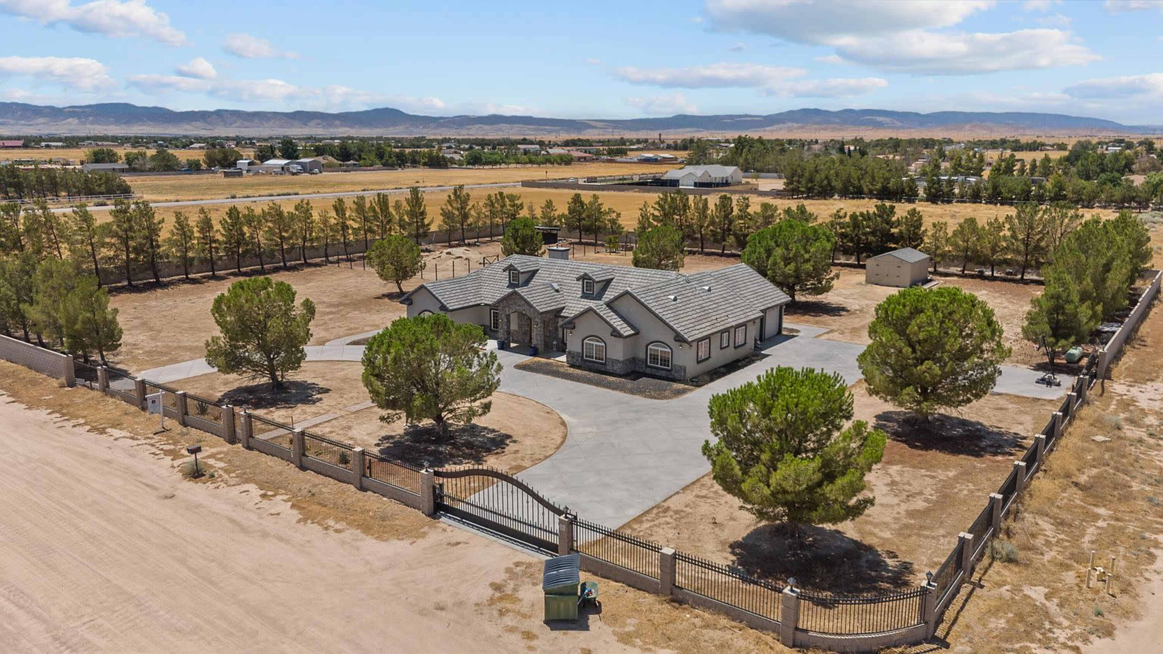 The image shows a large, single-story house surrounded by a fenced yard, with several trees and open land in the background.