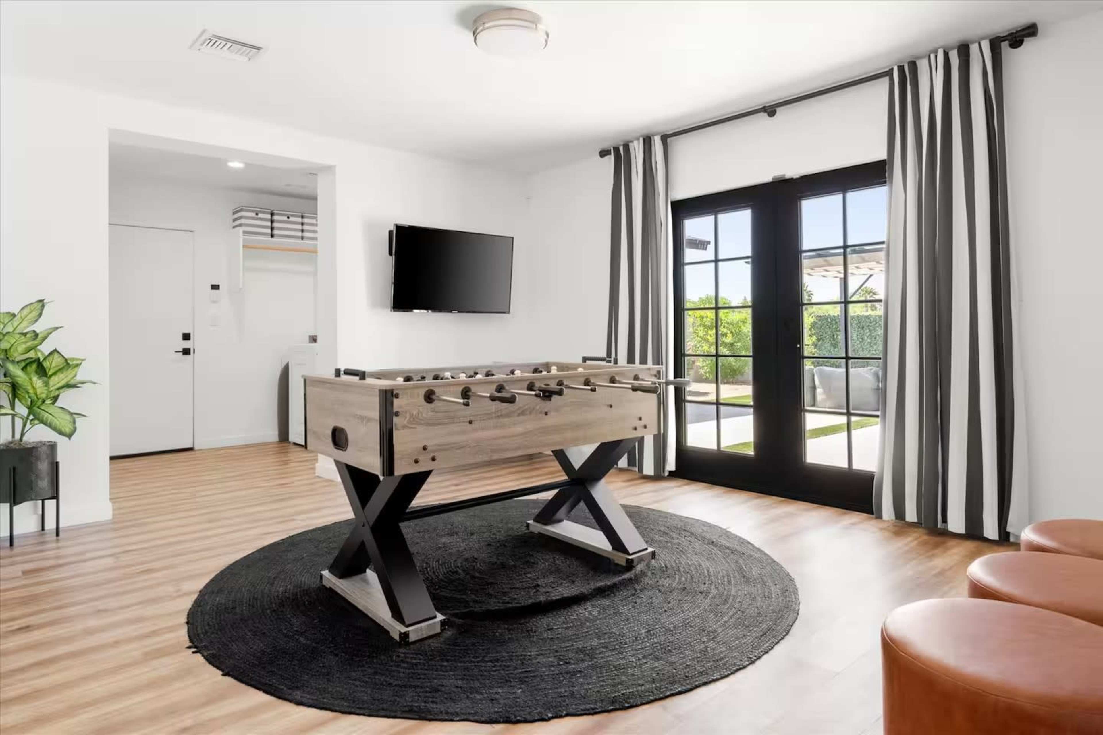 The image features a modern living space with a foosball table in the center, a television mounted on the wall, and large glass doors leading to an outdoor area, framed by striped curtains.