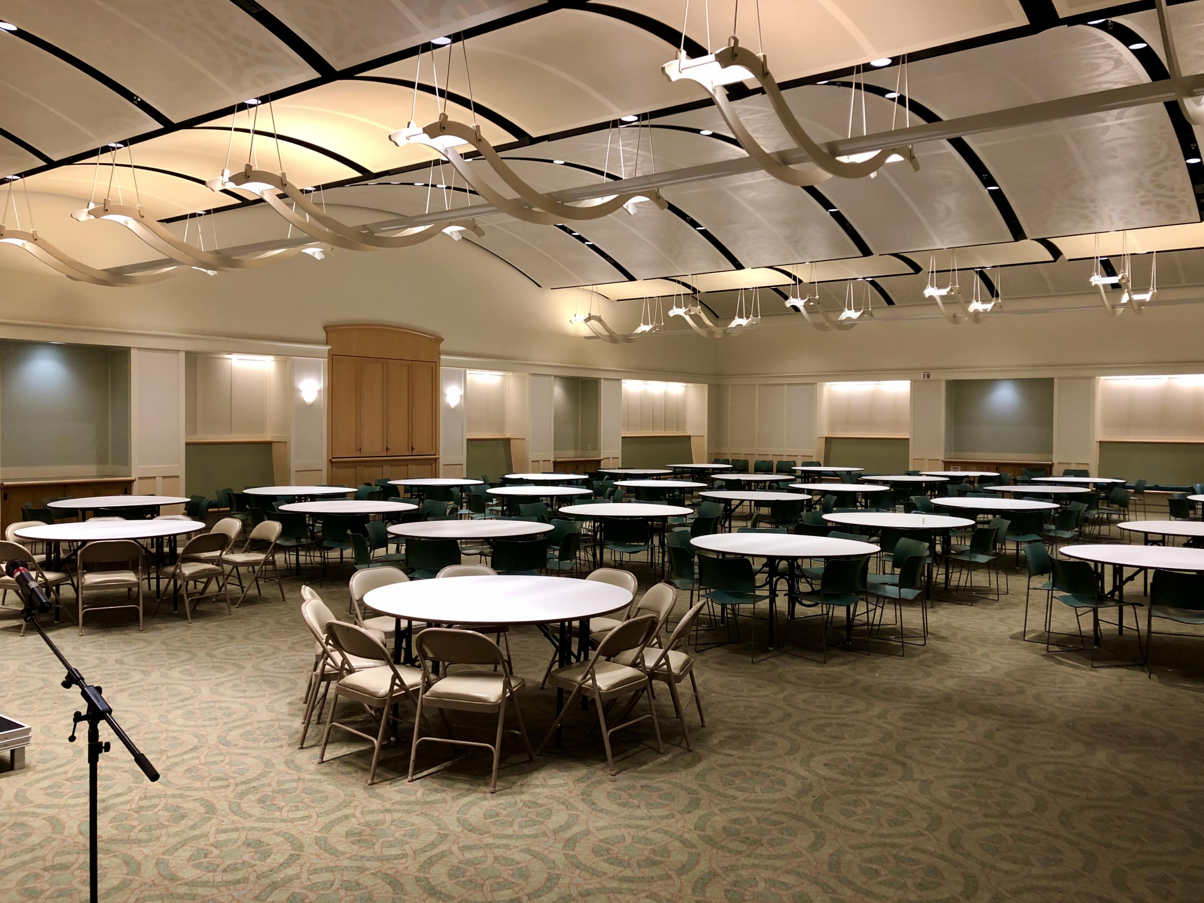 The image shows a spacious indoor venue arranged with multiple round tables and chairs, featuring a high ceiling with decorative lighting.