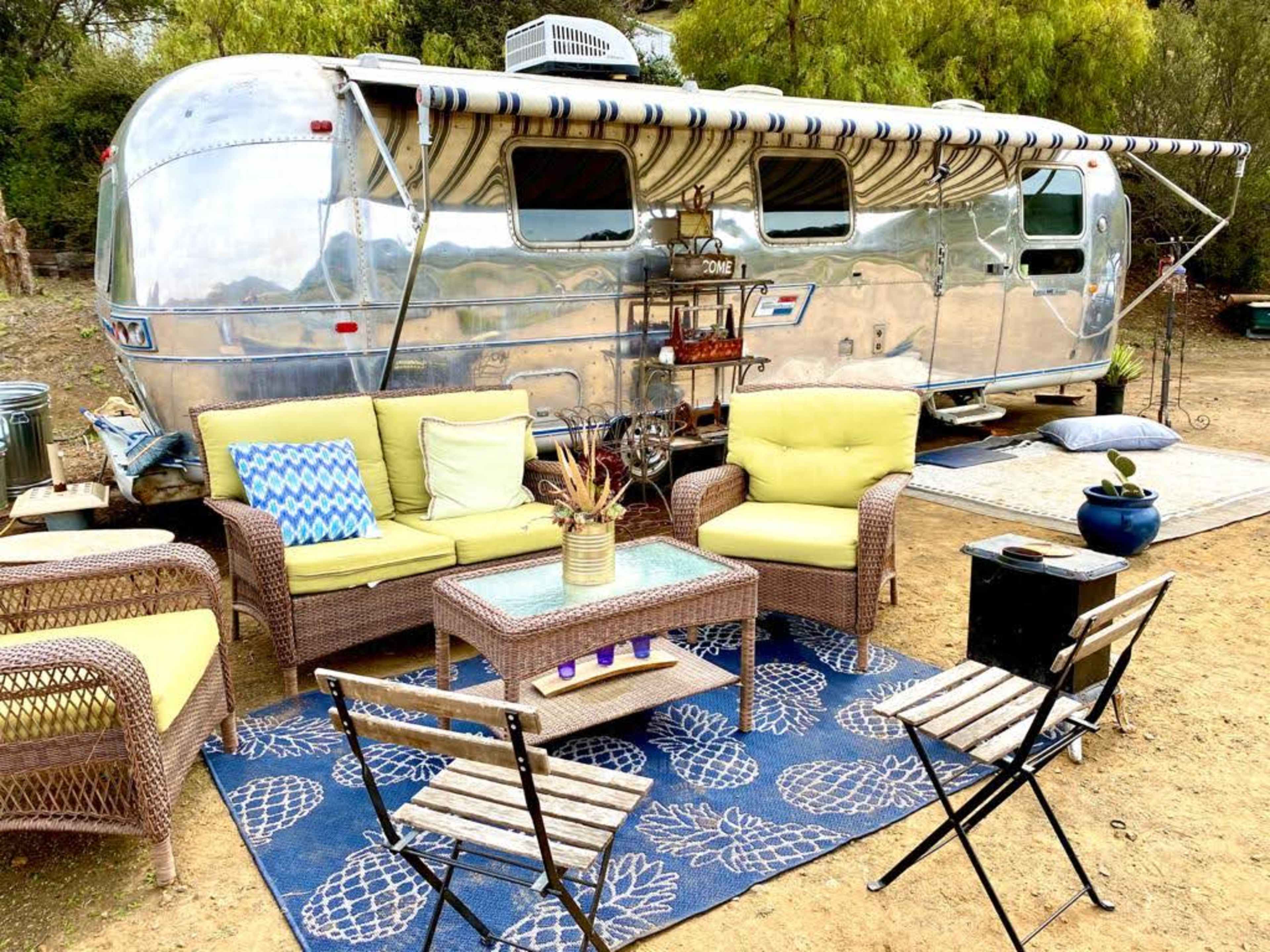 An Airstream trailer is parked next to a seating area with wicker chairs and a table surrounded by decorative items.