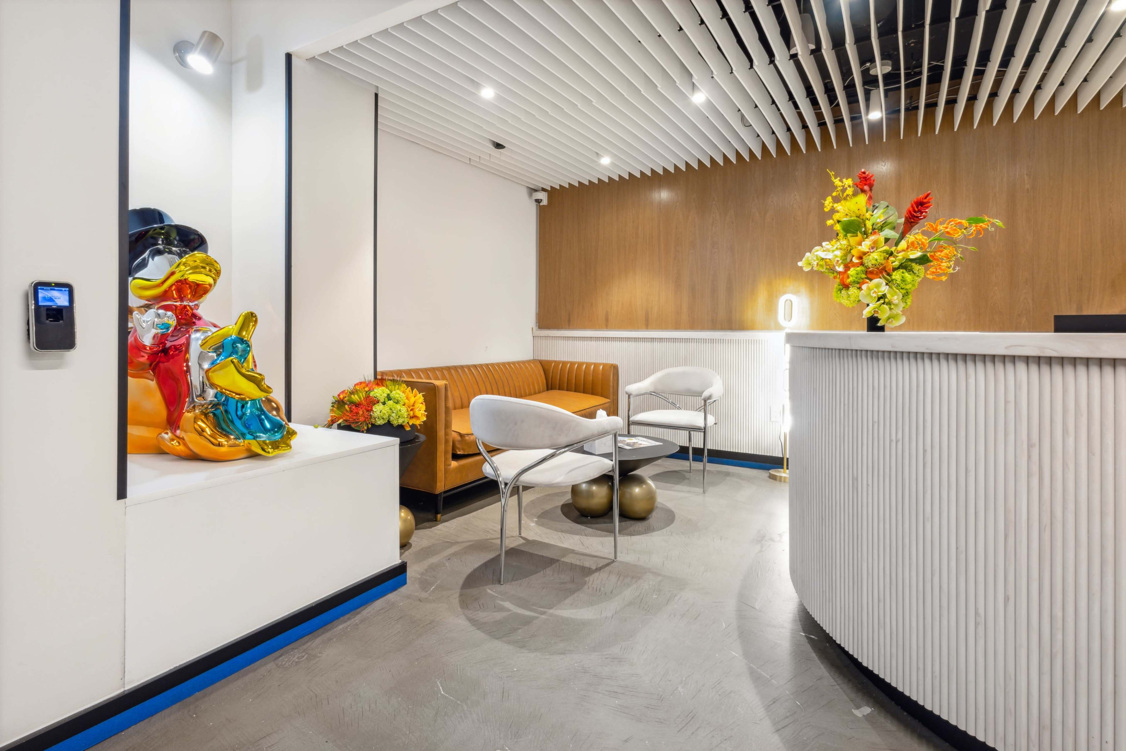 The image shows a modern reception area featuring a curved desk, a brown leather sofa, white chairs, colorful sculptures, and floral arrangements.