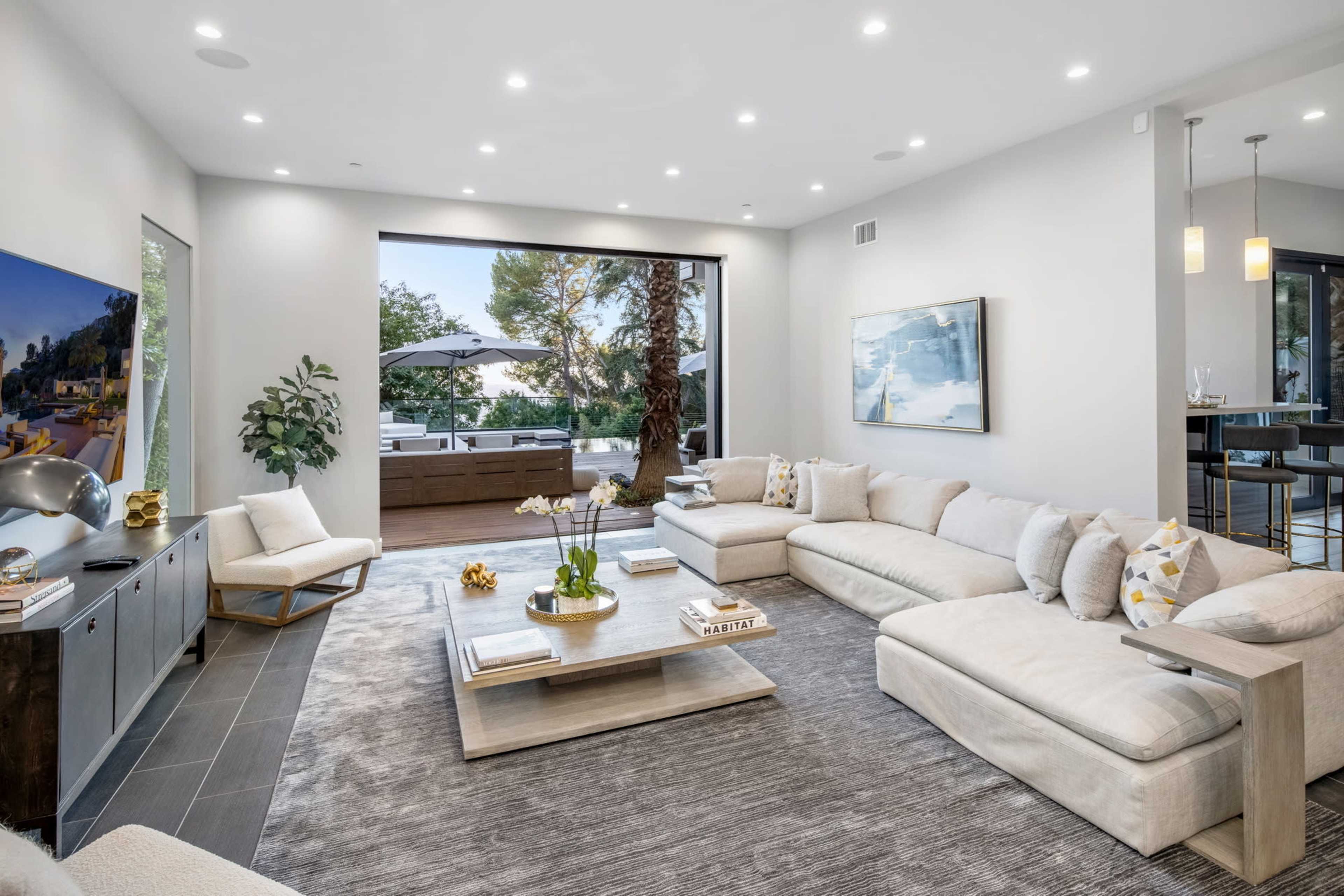 A modern living room features a large sectional sofa, a coffee table, and floor-to-ceiling windows opening to an outdoor patio with a pool.