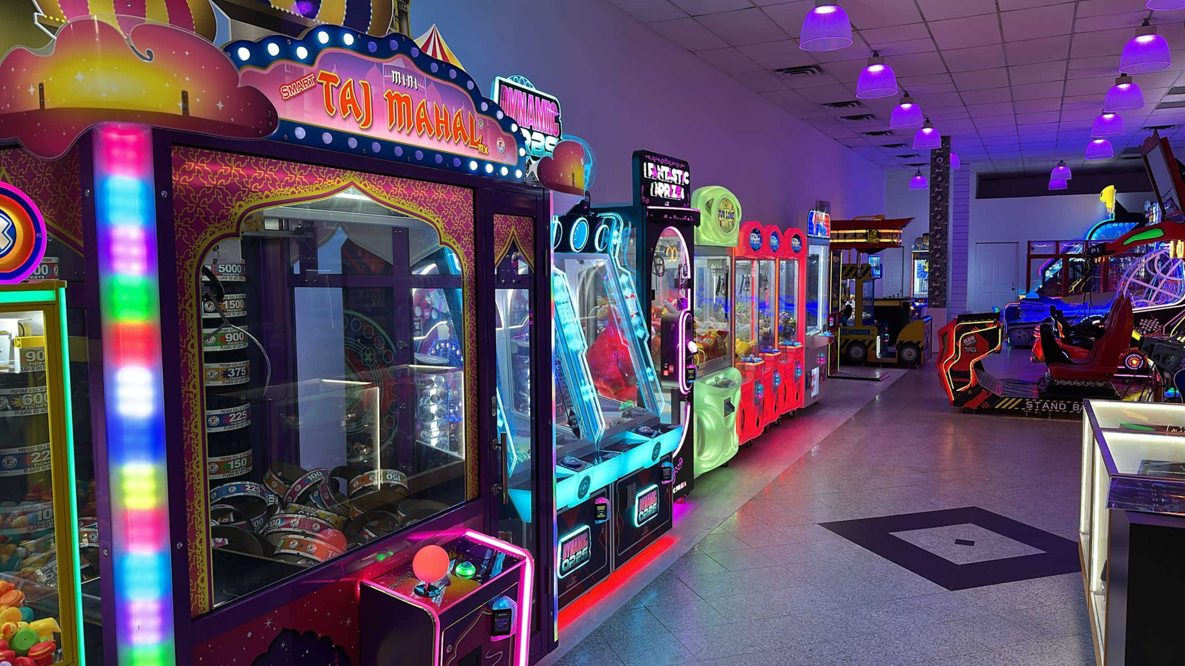 The image shows a brightly lit arcade featuring various colorful gaming machines and attractions along a corridor.