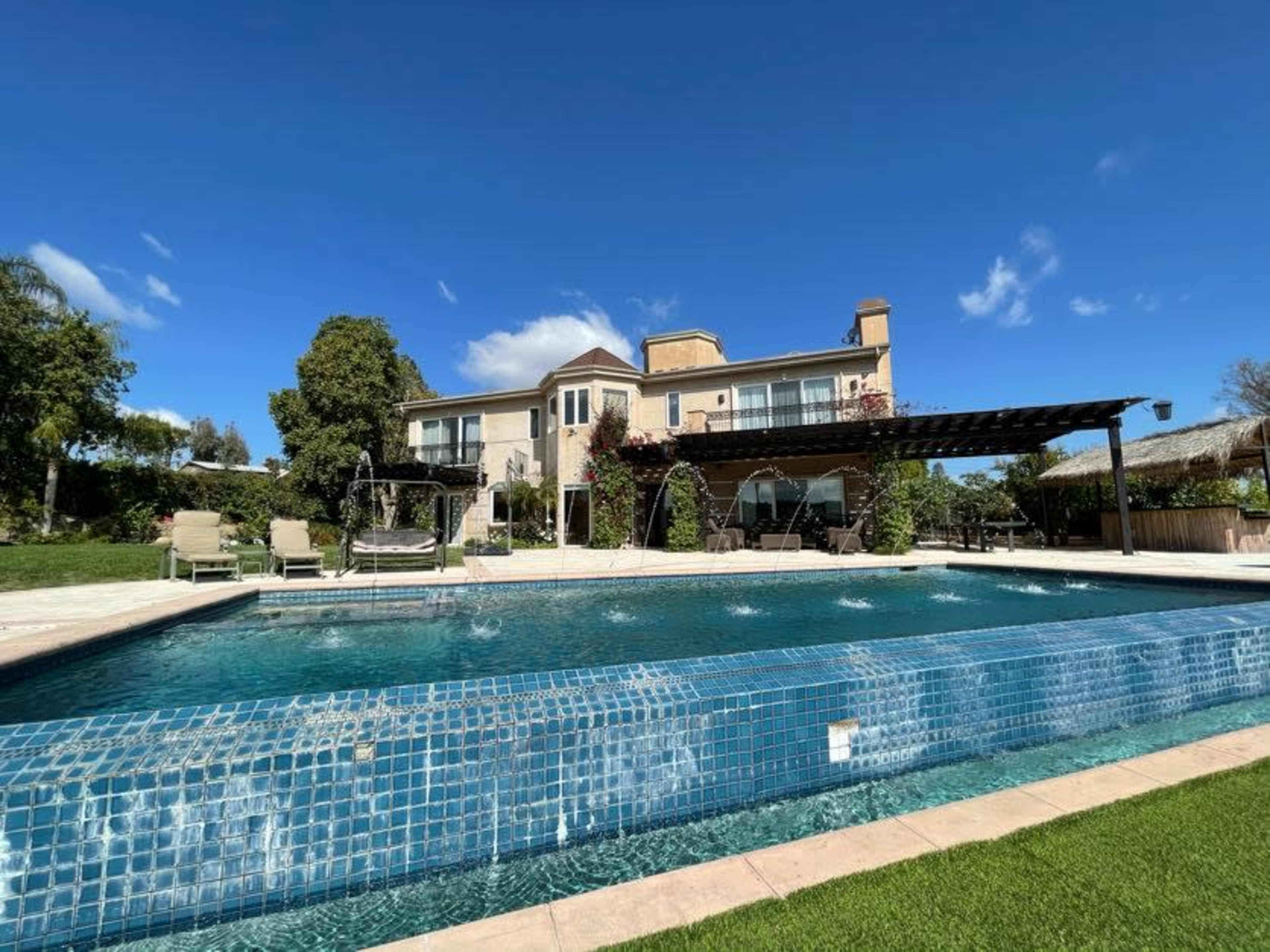 A spacious backyard features a swimming pool in front of a two-story house, surrounded by green grass and trees under a clear blue sky.